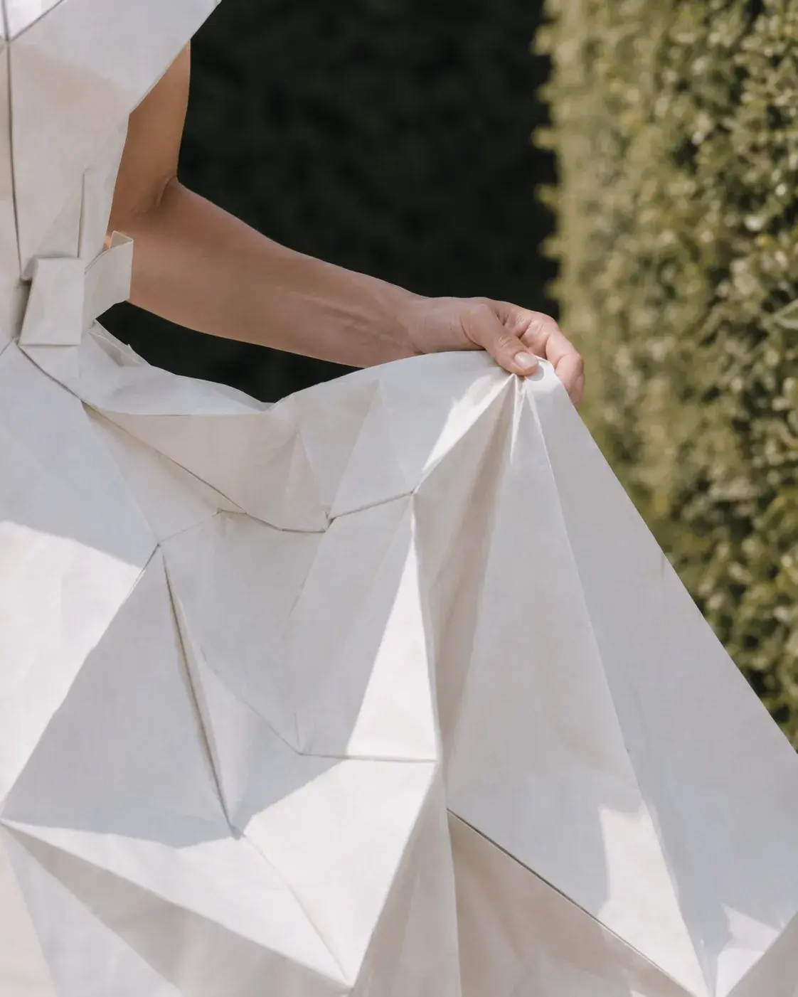 Gros plan main sur jupe origami géométrique – détail tissu facetté et haie verte en fond