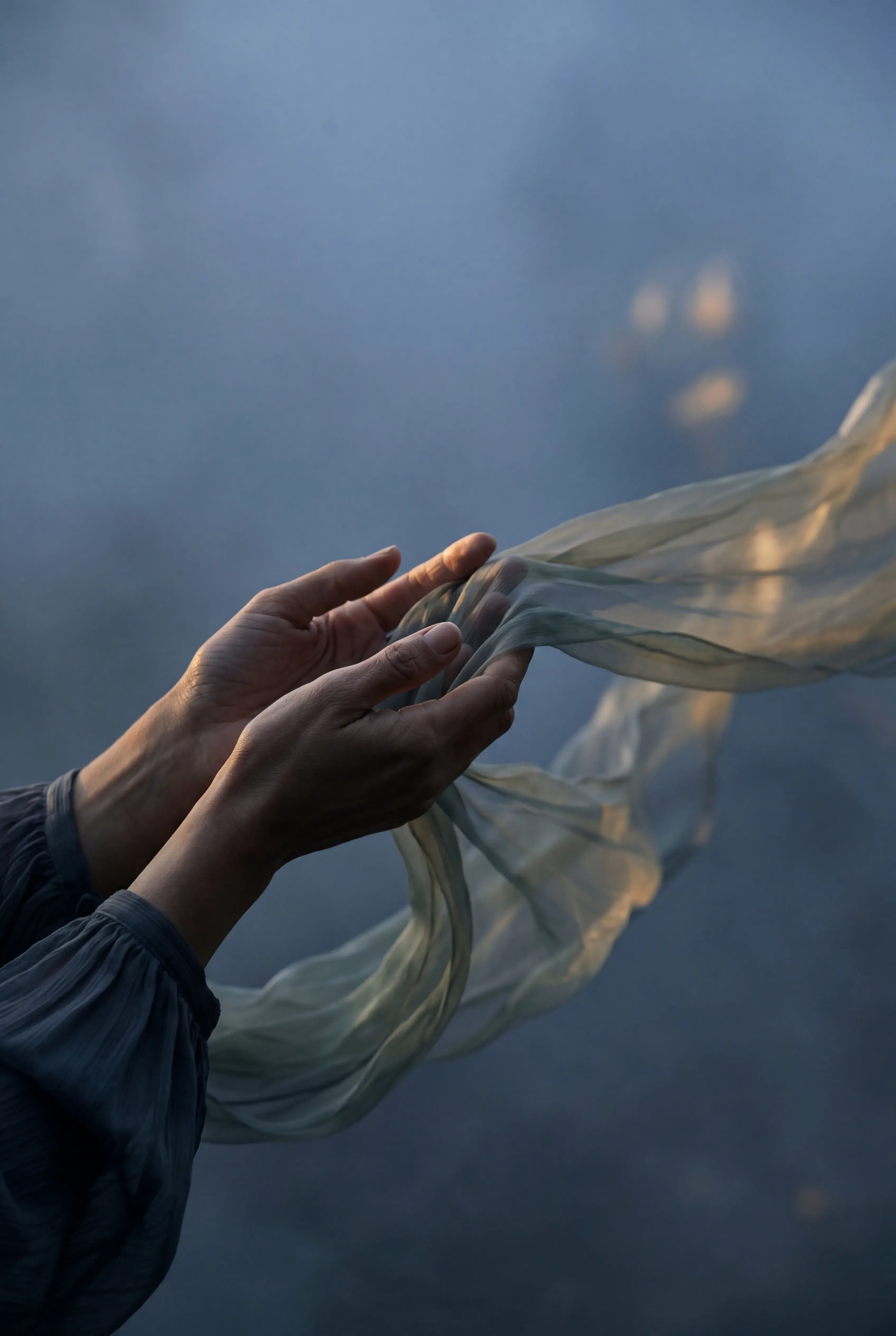 Gros plan mains tendues – voile gaze léger flottant depuis les doigts, lumière chaude, fond brumeux