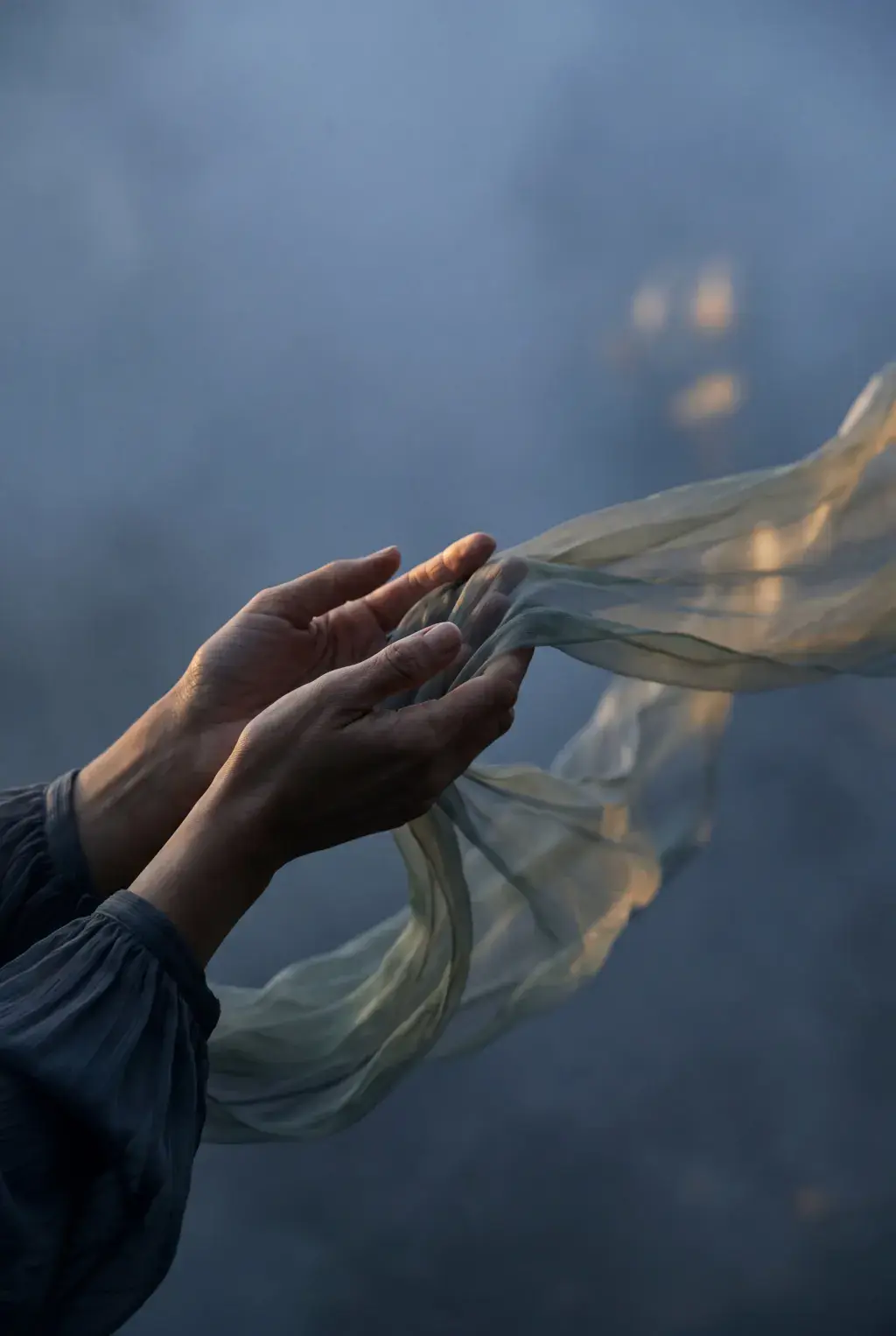 Gros plan mains tendues – voile gaze léger flottant depuis les doigts, lumière chaude, fond brumeux