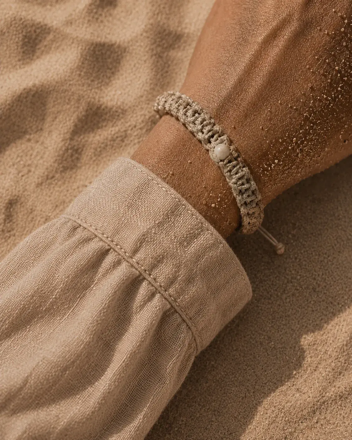 Gros plan poignet avec bracelet macramé beige – perle blanche, sable doré sur peau, manche lin