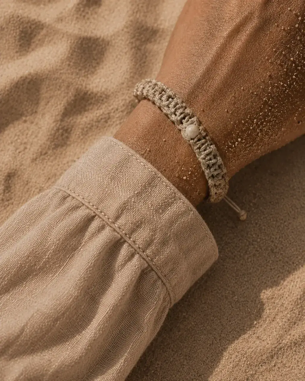 Gros plan poignet avec bracelet macramé beige – perle blanche, sable doré sur peau, manche lin