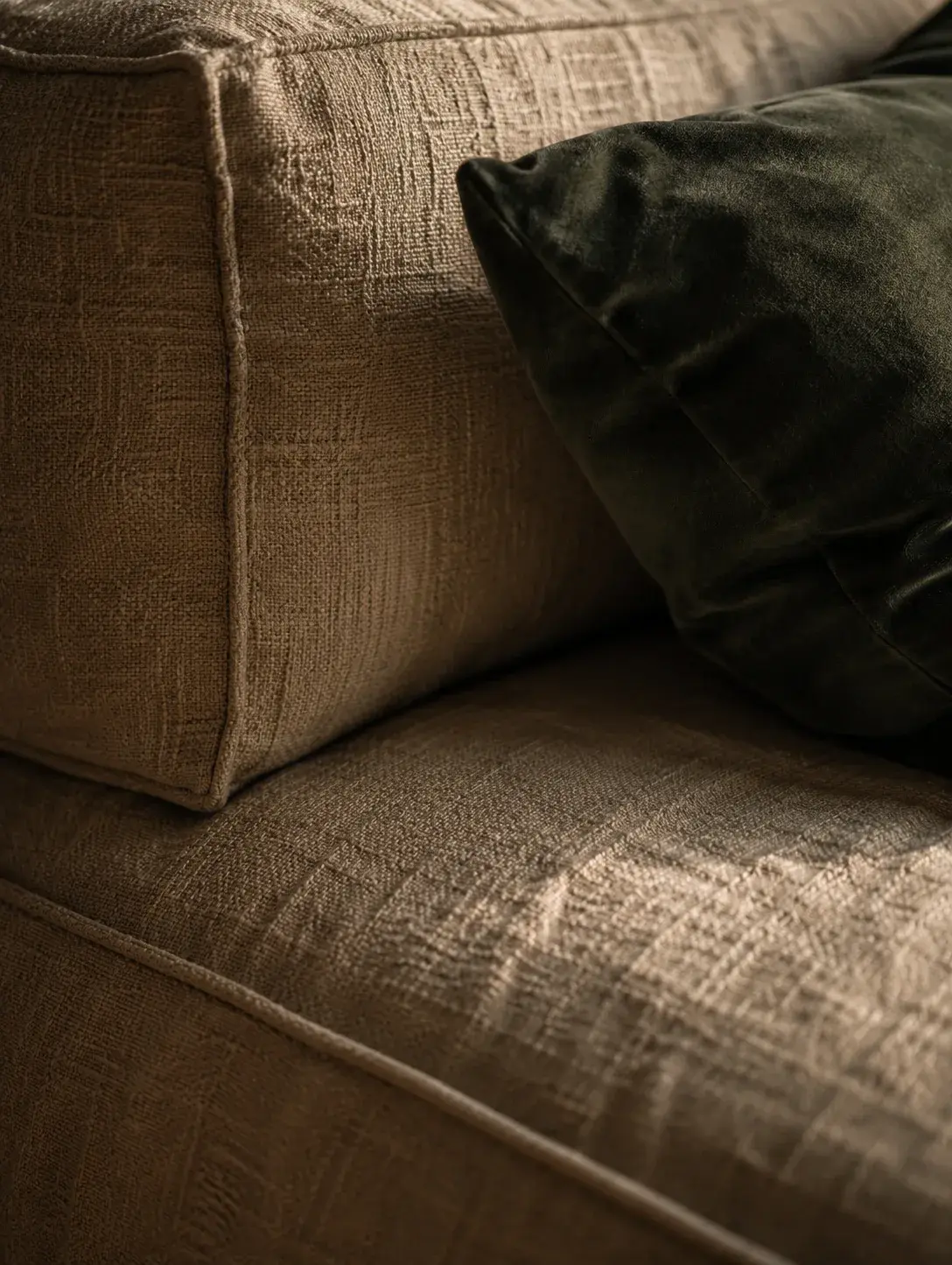 Gros plan sur accoudoir de canapé en lin texturé beige avec coussin velours vert foncé, lumière rasante