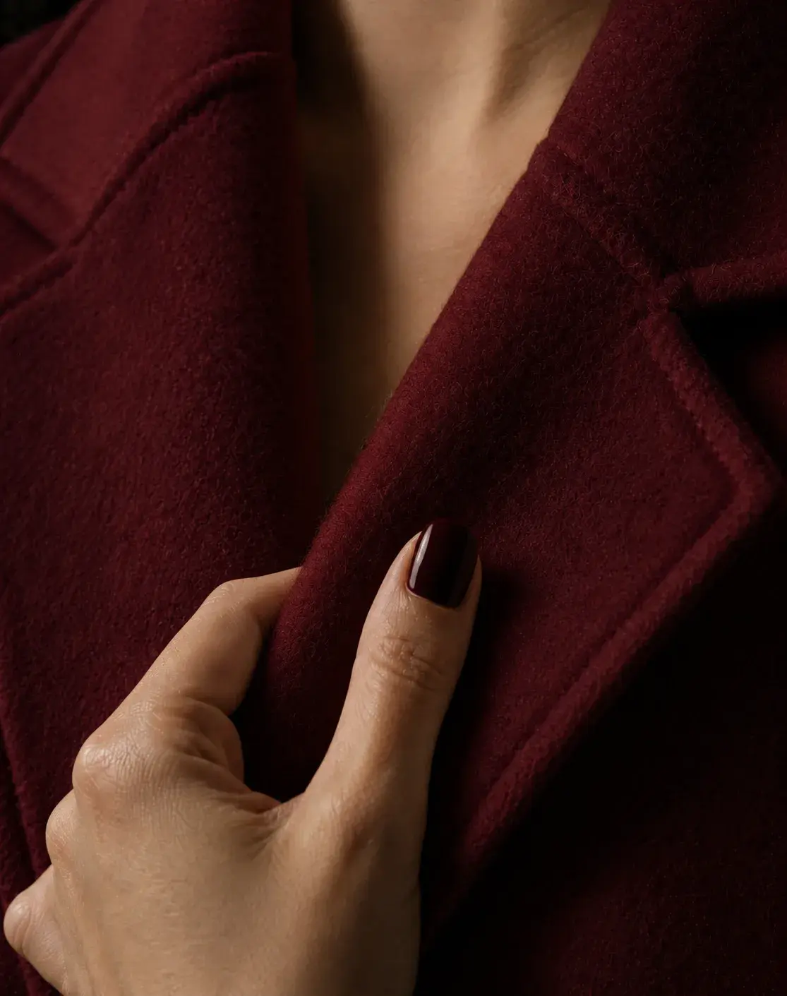 Gros plan sur main féminine (ongles bordeaux) tenant le revers d'un manteau en laine bordeaux, peau nue entrevue