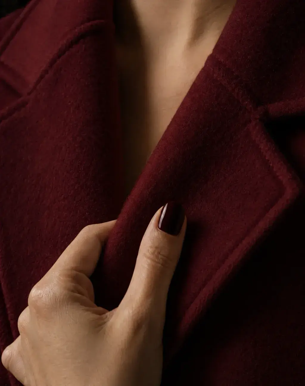 Gros plan sur main féminine (ongles bordeaux) tenant le revers d'un manteau en laine bordeaux, peau nue entrevue