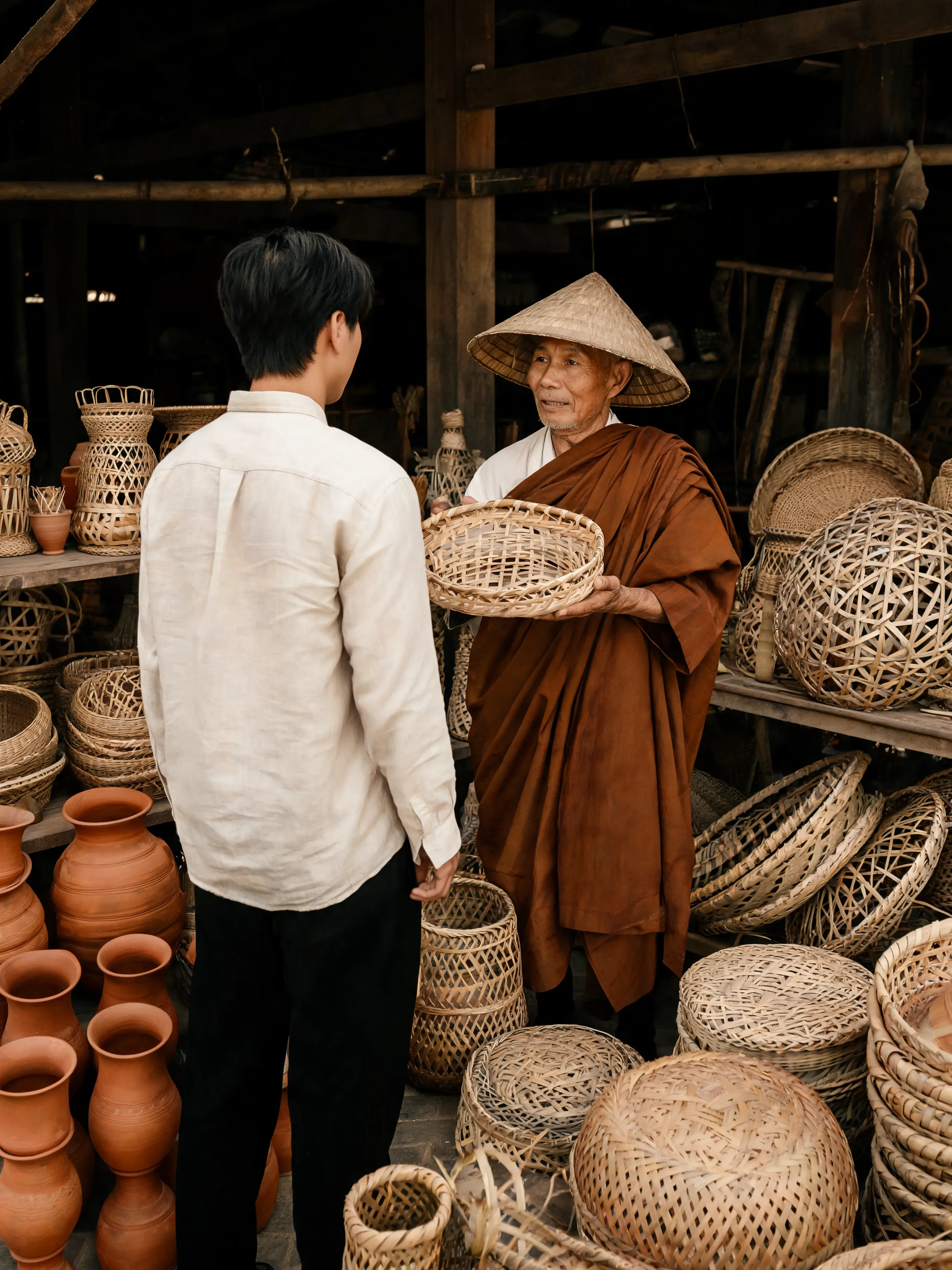 Homme de dos en tenue lin blanc face à un moine-artisan senior en chapeau conique présentant un plateau tressé dans une boutique pleine de paniers et céramique