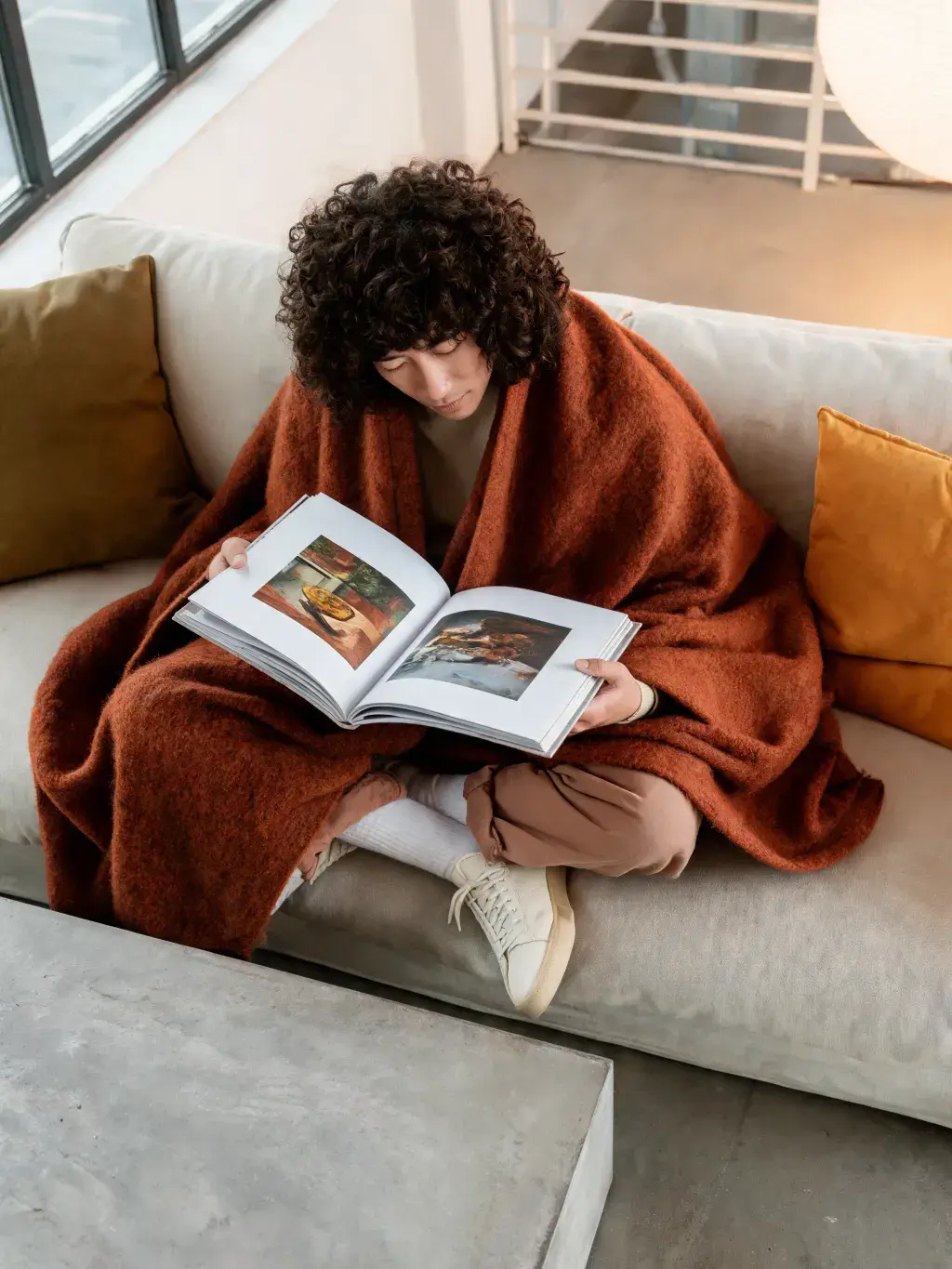 Homme enroulé plaid terracotta lisant livre d'art – canapé, coussins, lumière naturelle, loft