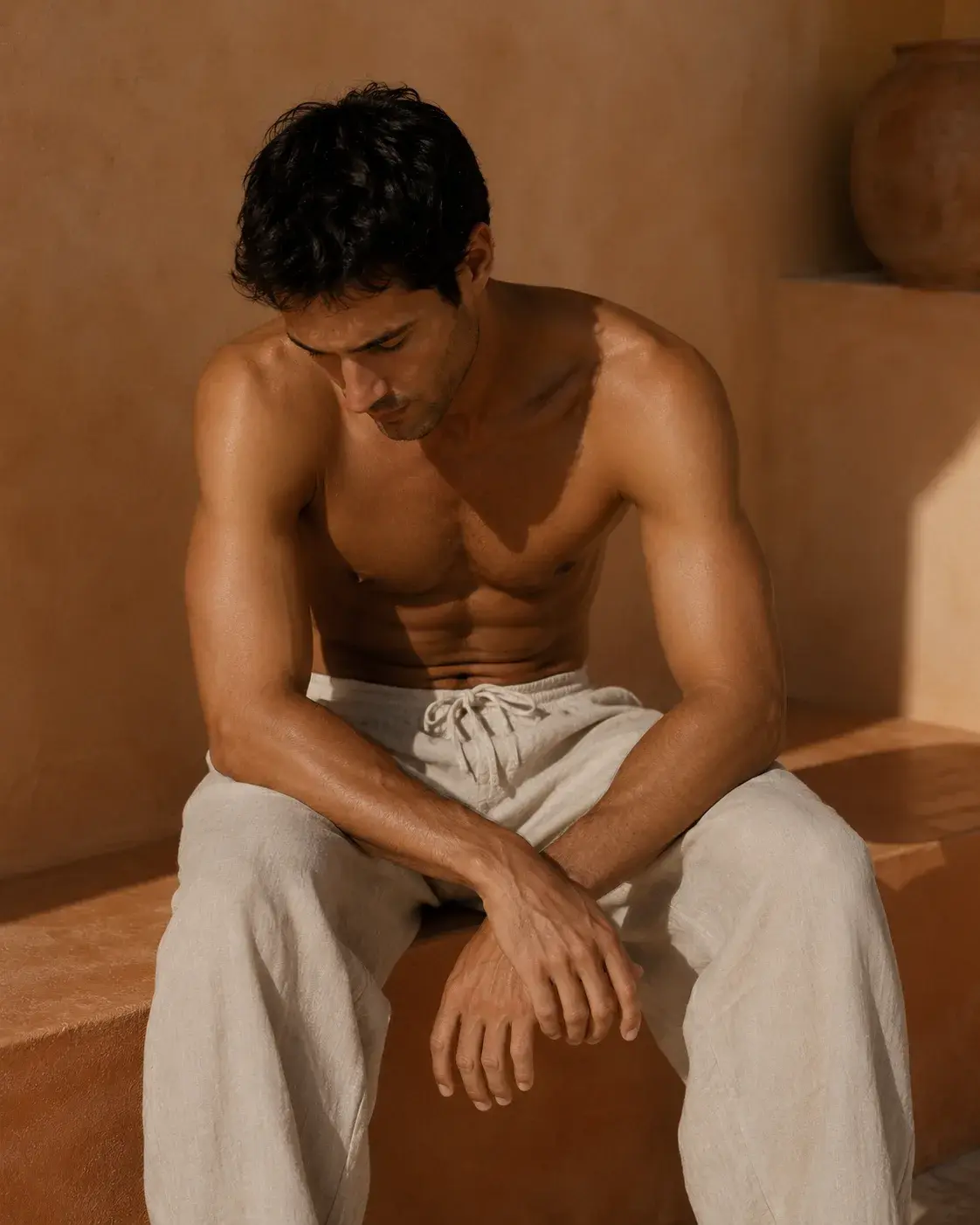 Homme torse nu assis sur rebord de pierre – pantalon lin beige, mur ocre désertique, lumière chaude