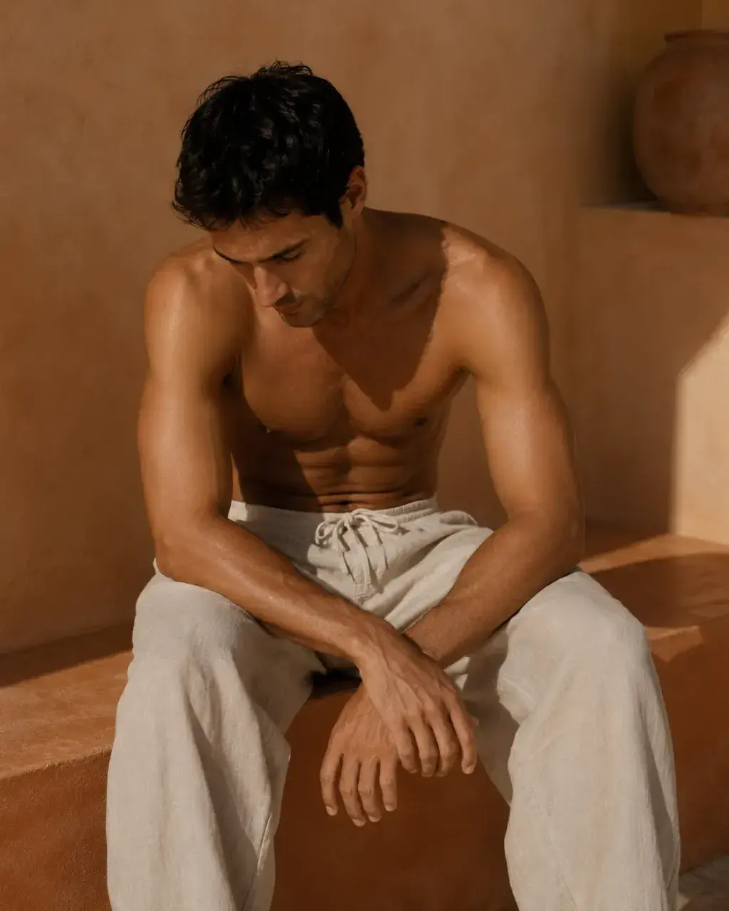 Homme torse nu assis sur rebord de pierre – pantalon lin beige, mur ocre désertique, lumière chaude