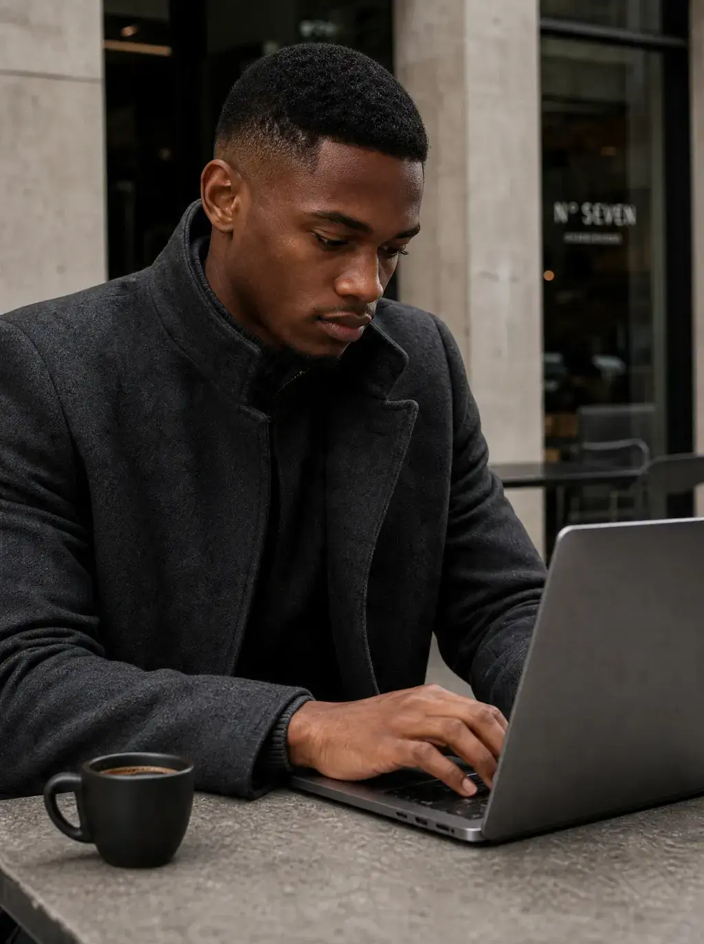 Homme travaillant sur laptop – table café extérieure, mug noir, manteau gris, façade urbaine
