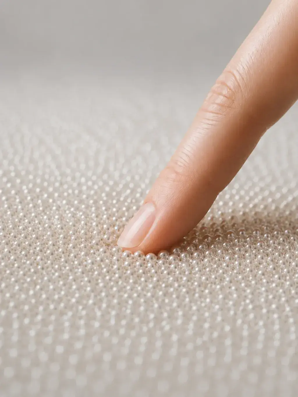 Index appuyant sur un tapis dense de micro-perles nacrées, geste tactile en gros plan