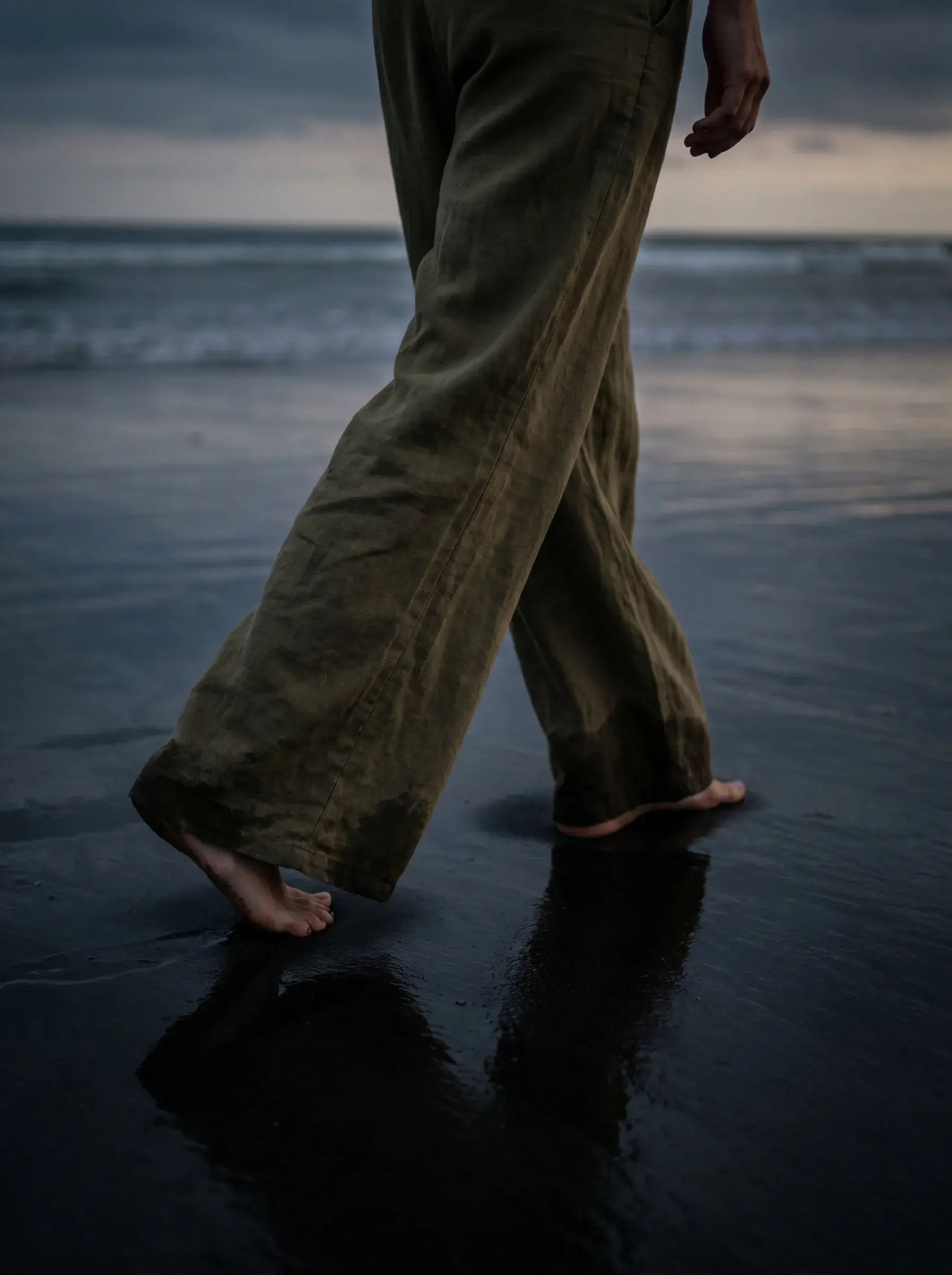 Jambes en pantalon lin kaki marchant pieds nus sur sable noir mouillé en bord de mer