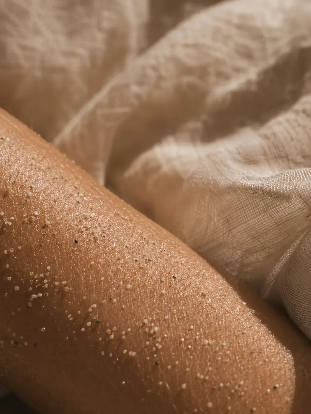 Macro épaule bronzée avec gommage sel – grains blancs sur peau, tissu mousseline, lumière sombre