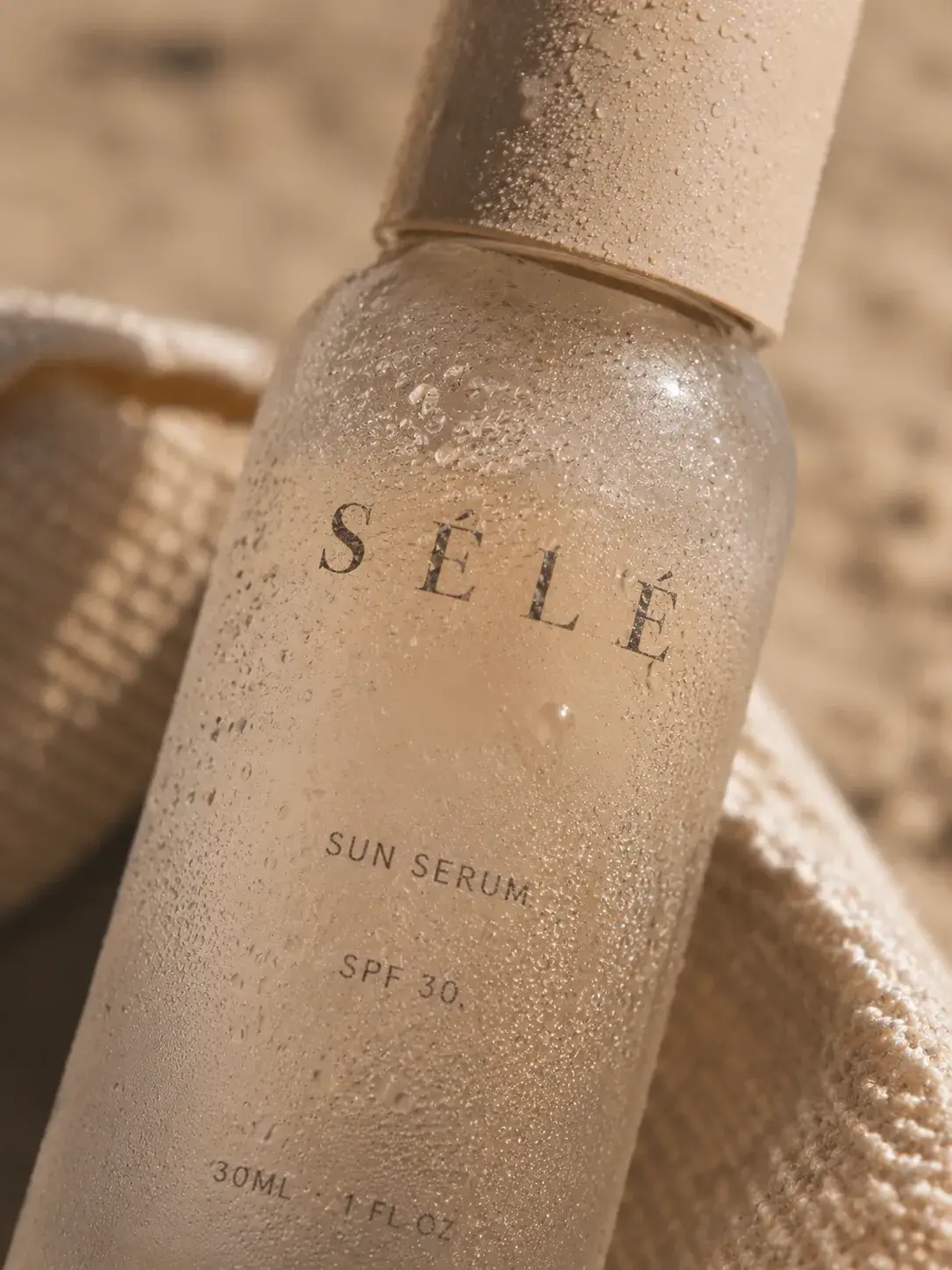 Macro flacon sérum verre couvert de gouttelettes et sable – tissu raphia, lumière chaude estivale