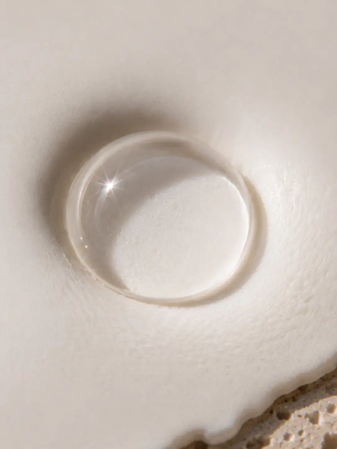 Macro goutte de sérum translucide avec reflet étoilé – crème blanche, fond plâtre, lumière douce
