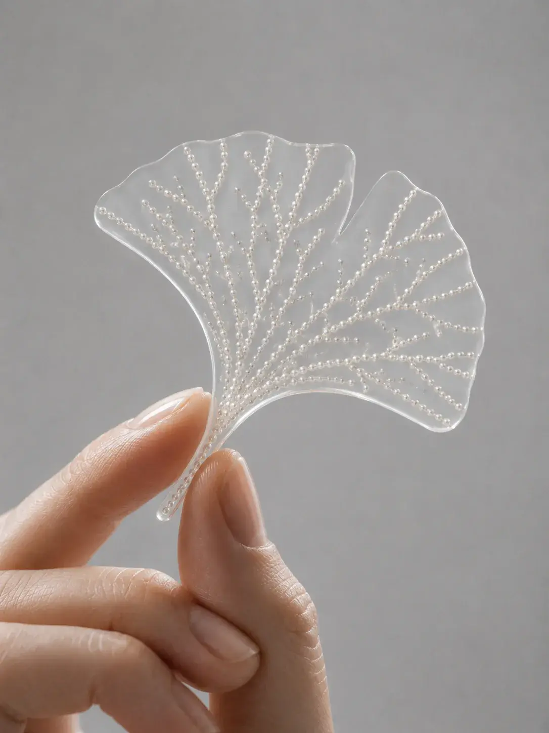Main à trois doigts tenant une feuille de ginkgo translucide aux nervures incrustées de perles, fond gris