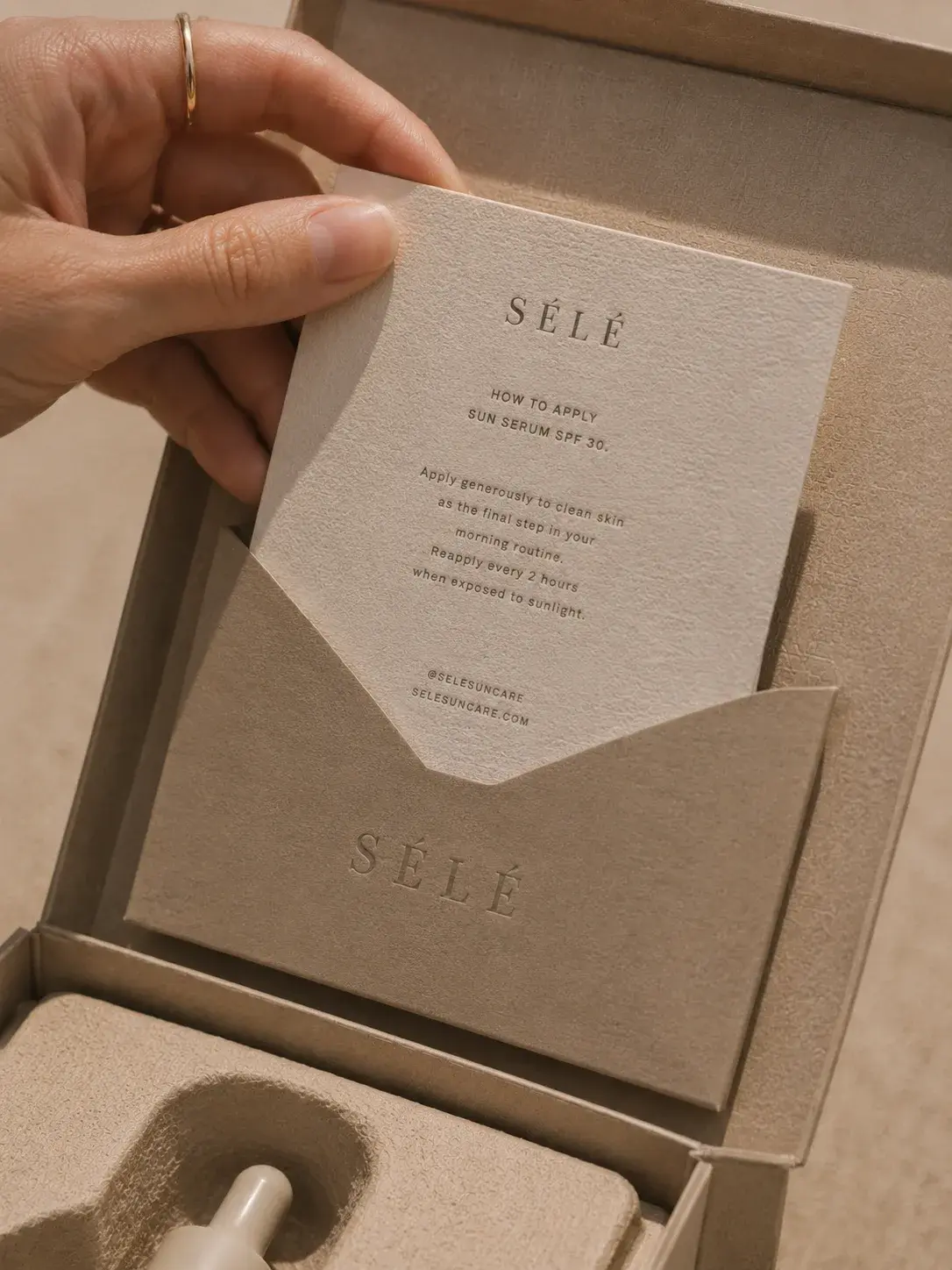Main avec bague dorée soulevant une carte d'instructions dans coffret – flacon sérum, packaging