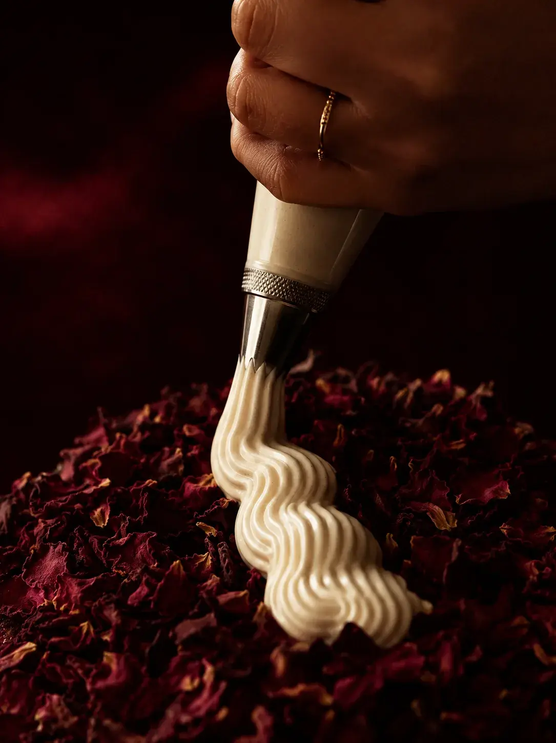 Main avec bague dorée utilisant une poche à douille sur un lit de pétales de rose séchées rouges, crème fouettée en spirale