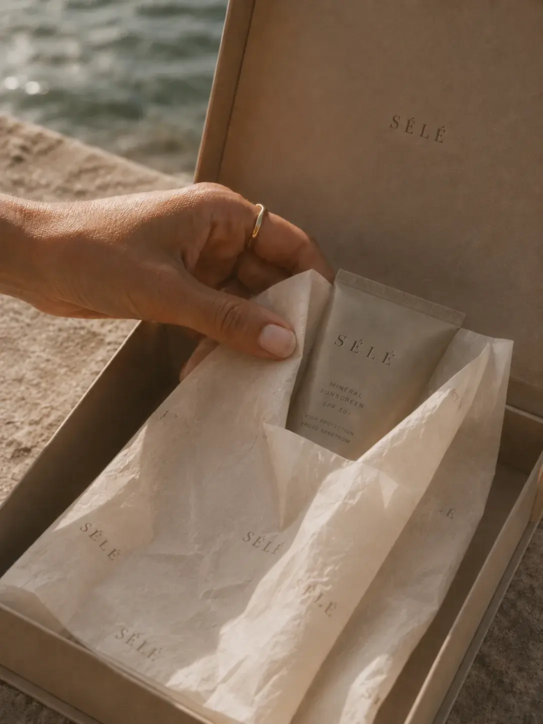 Main avec bague plongeant dans coffret kraft – tube soin, papier de soie, bord de mer en fond