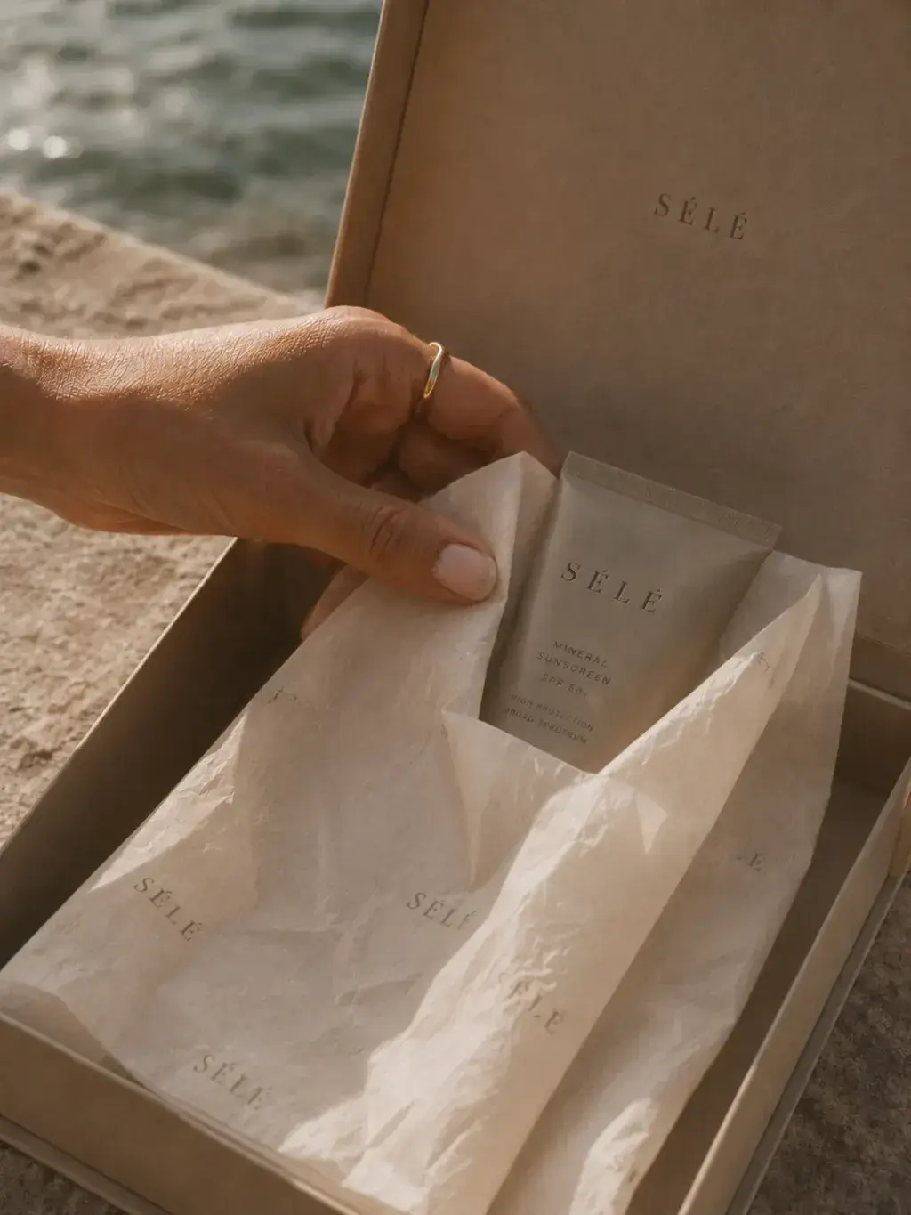 Main avec bague plongeant dans coffret kraft – tube soin, papier de soie, bord de mer en fond