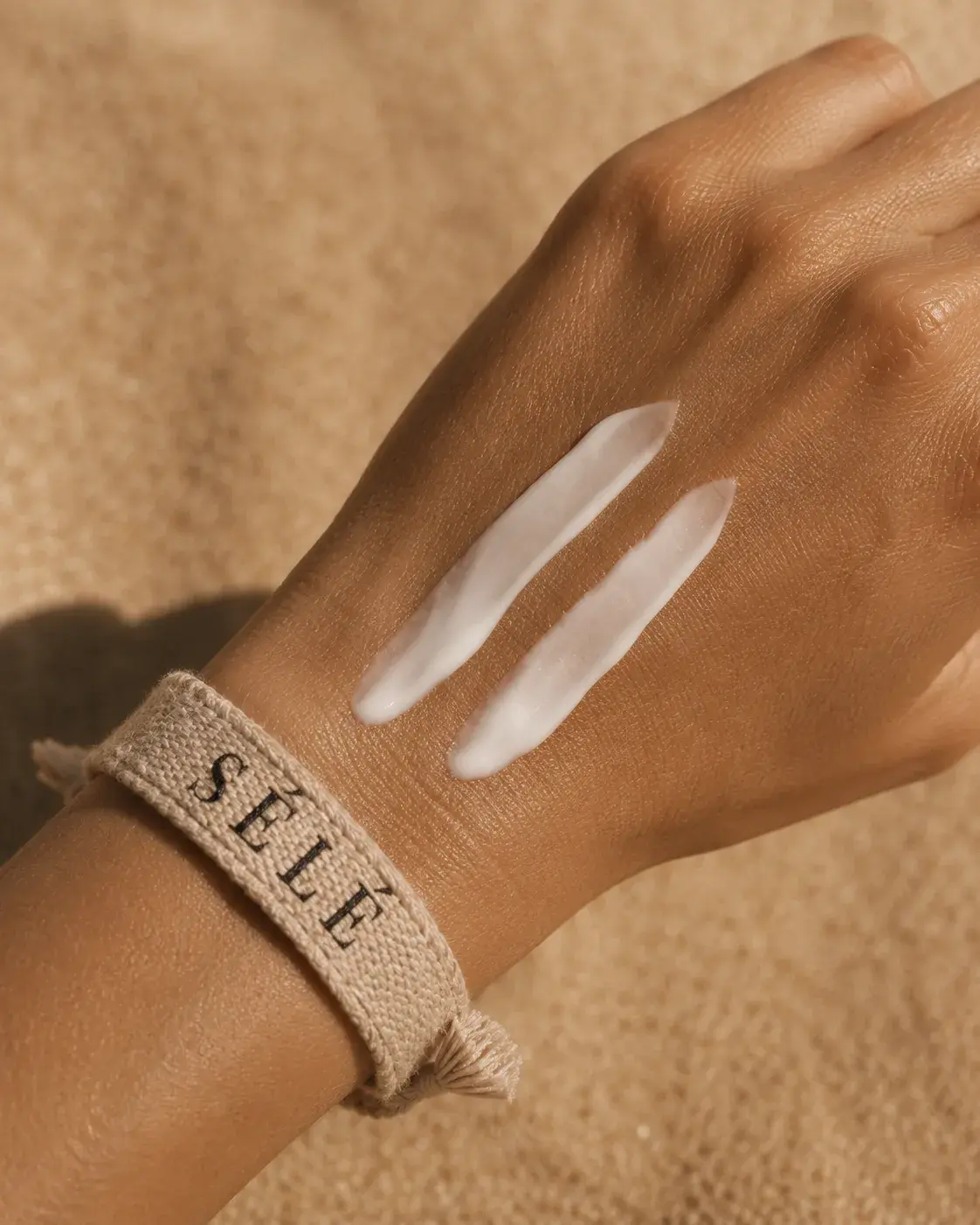 Main bronzée avec deux traces de crème blanche – bracelet tissu jute beige, fond sable
