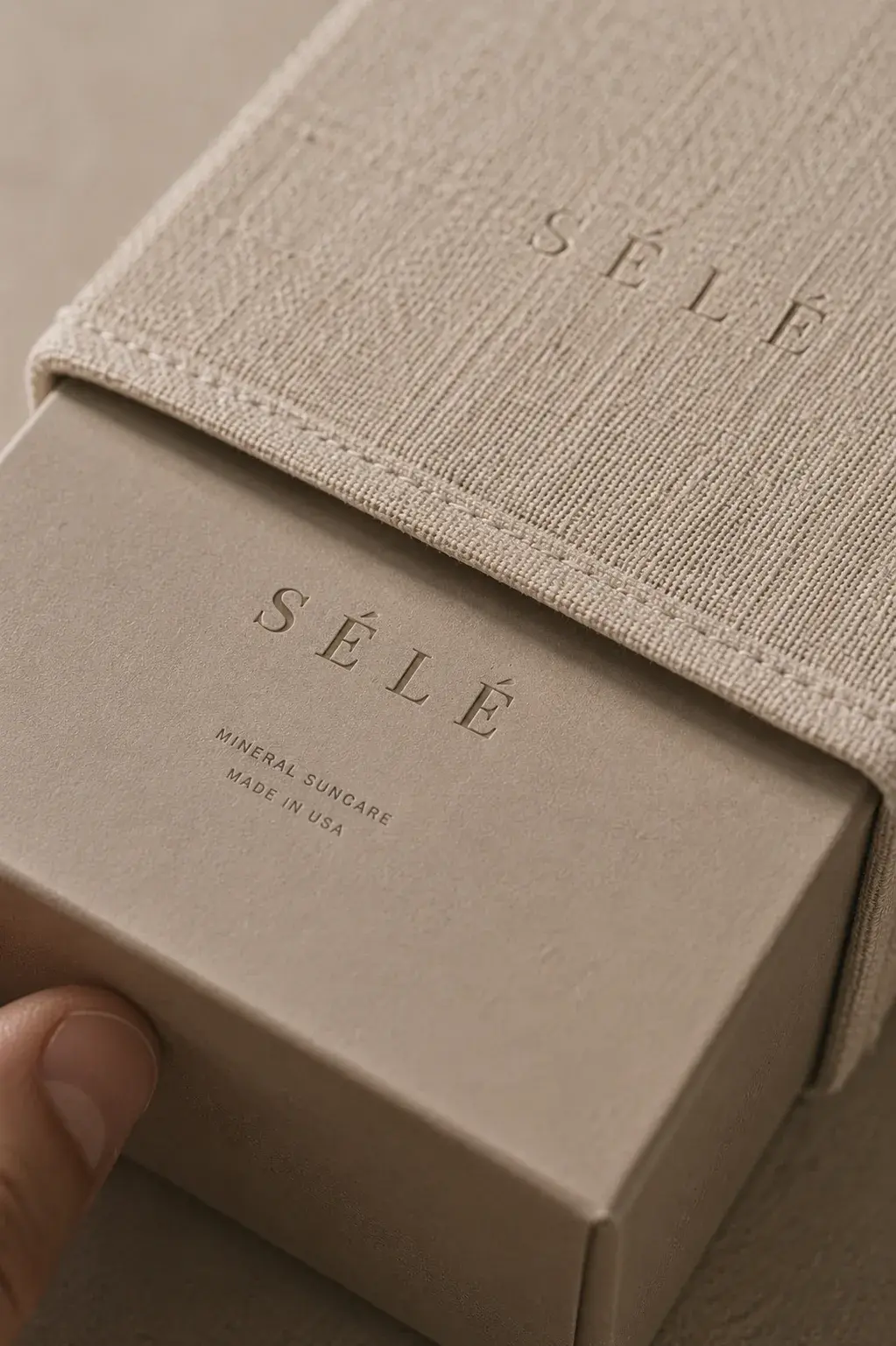Main extrayant une boîte kraft d'un coffret tissu texturé beige – packaging luxe, fond neutre