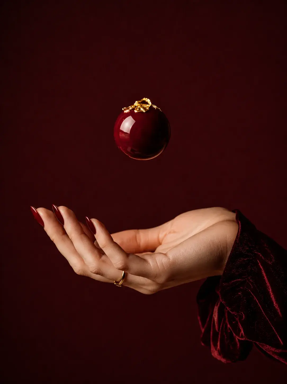 Main féminine avec bague dorée, entremets sphérique bordeaux en lévitation – fond bordeaux
