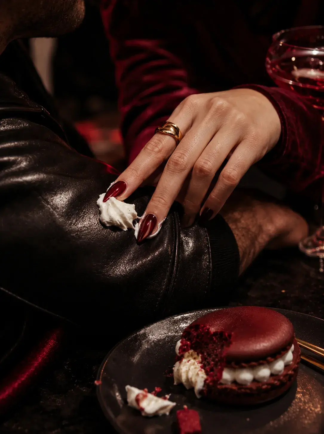 Main féminine avec macaron bordeaux sur botte cuir noire – ongles rouges, bague or, mood sombre