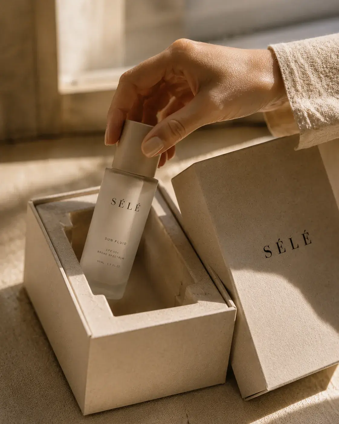 Main féminine saisissant un flacon sérum dans sa boîte – coffret kraft, papier de soie, unboxing
