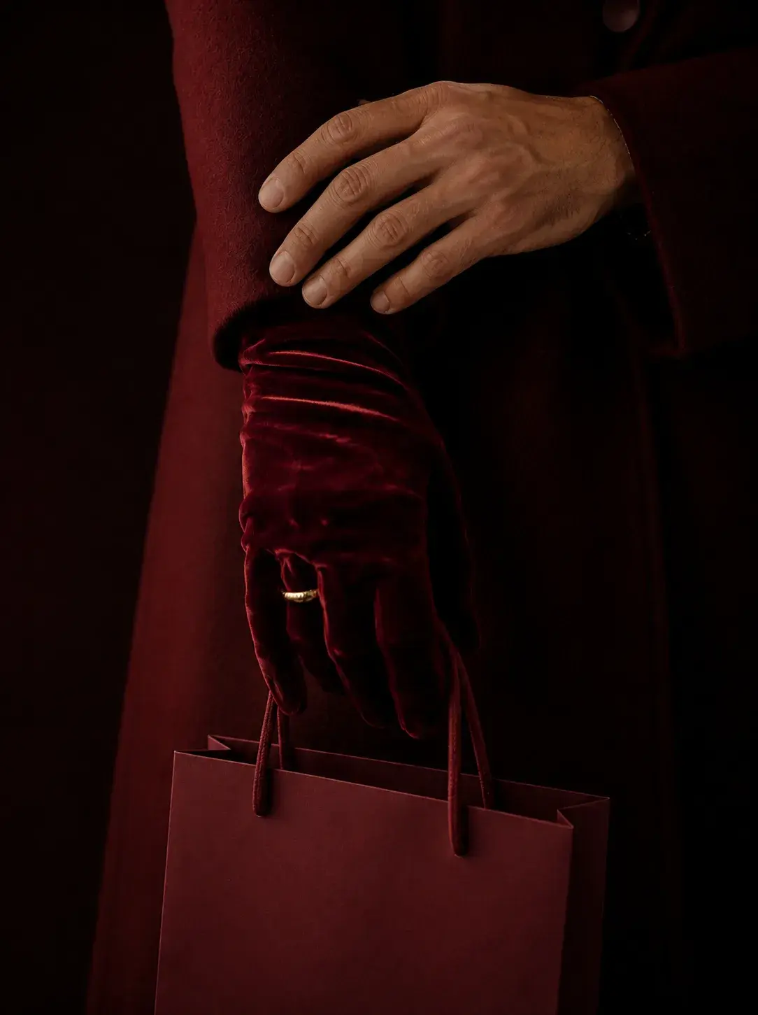 Main masculine posée sur main en gant de velours bordeaux tenant un sac shopping bordeaux, manteau bordeaux en fond