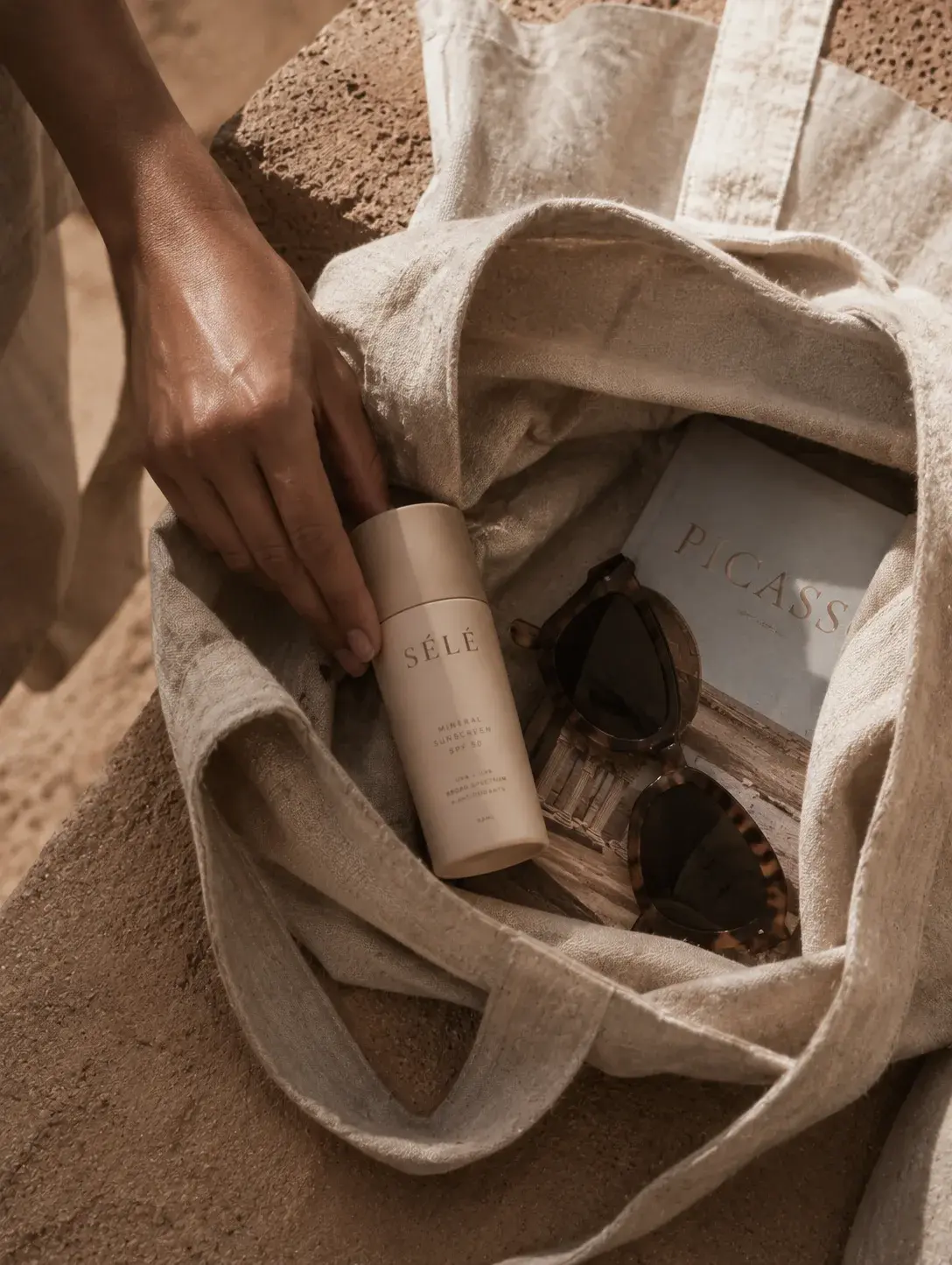 Main plongeant dans tote bag lin – flacon soin, lunettes écaille, livre, sable désertique