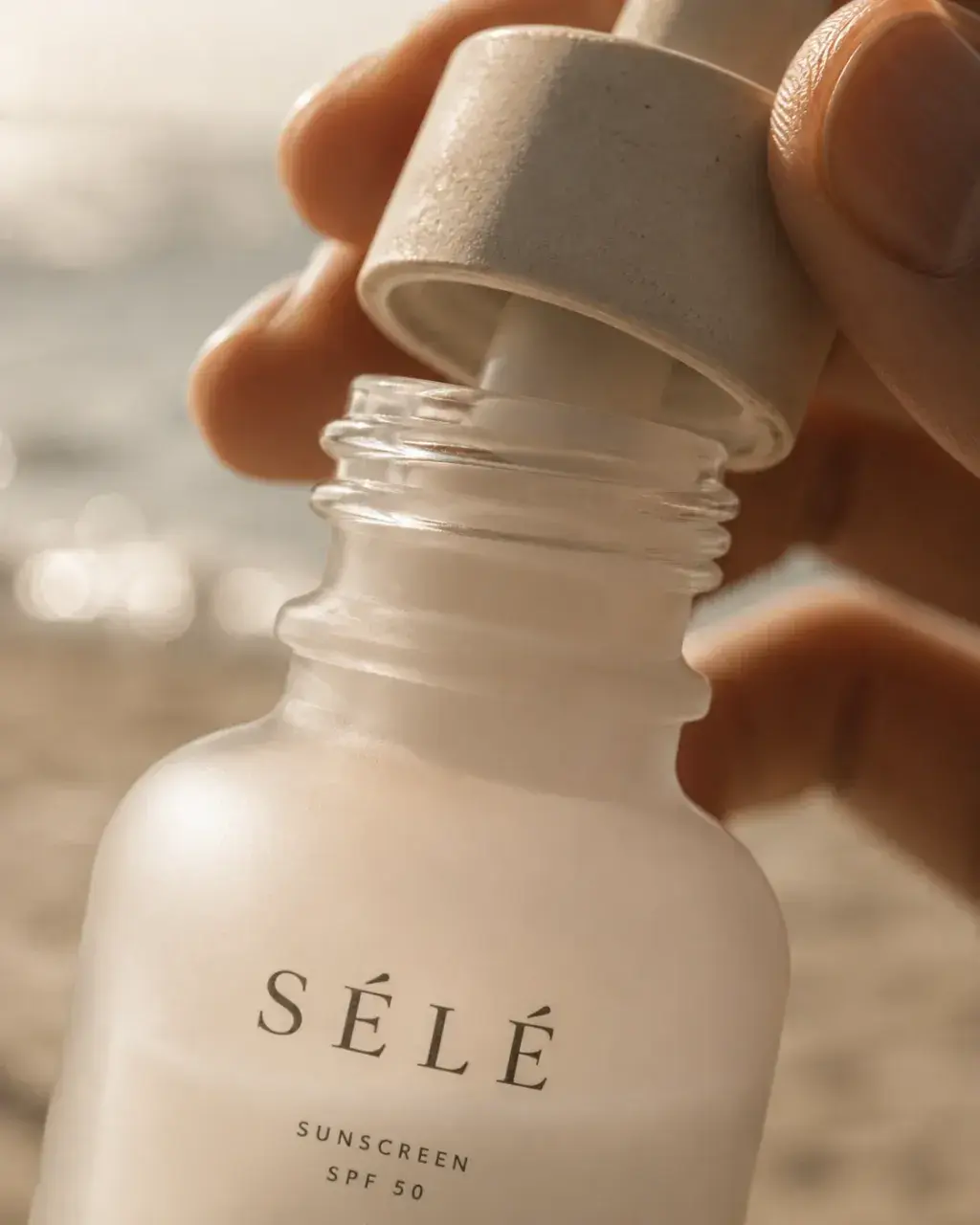 Main soulevant le bouchon d'un flacon de crème solaire en verre – mer en flou