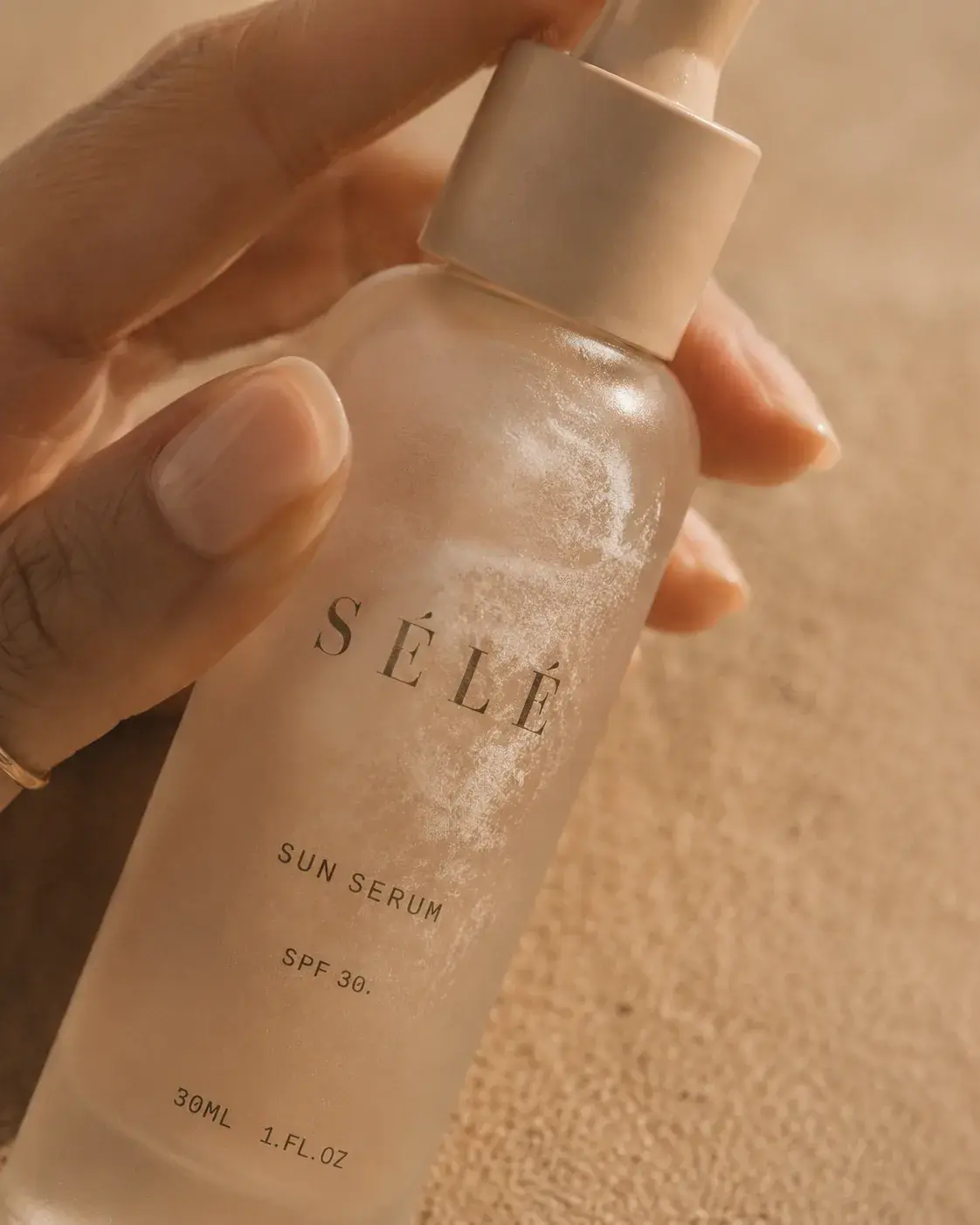 Main tenant flacon sérum pipette dans le sable – ongles naturels, fond plage, lumière chaude