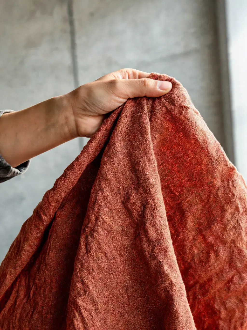 Main tenant tissu lin terracotta froissé – texture naturelle, fond béton gris, studio atelier