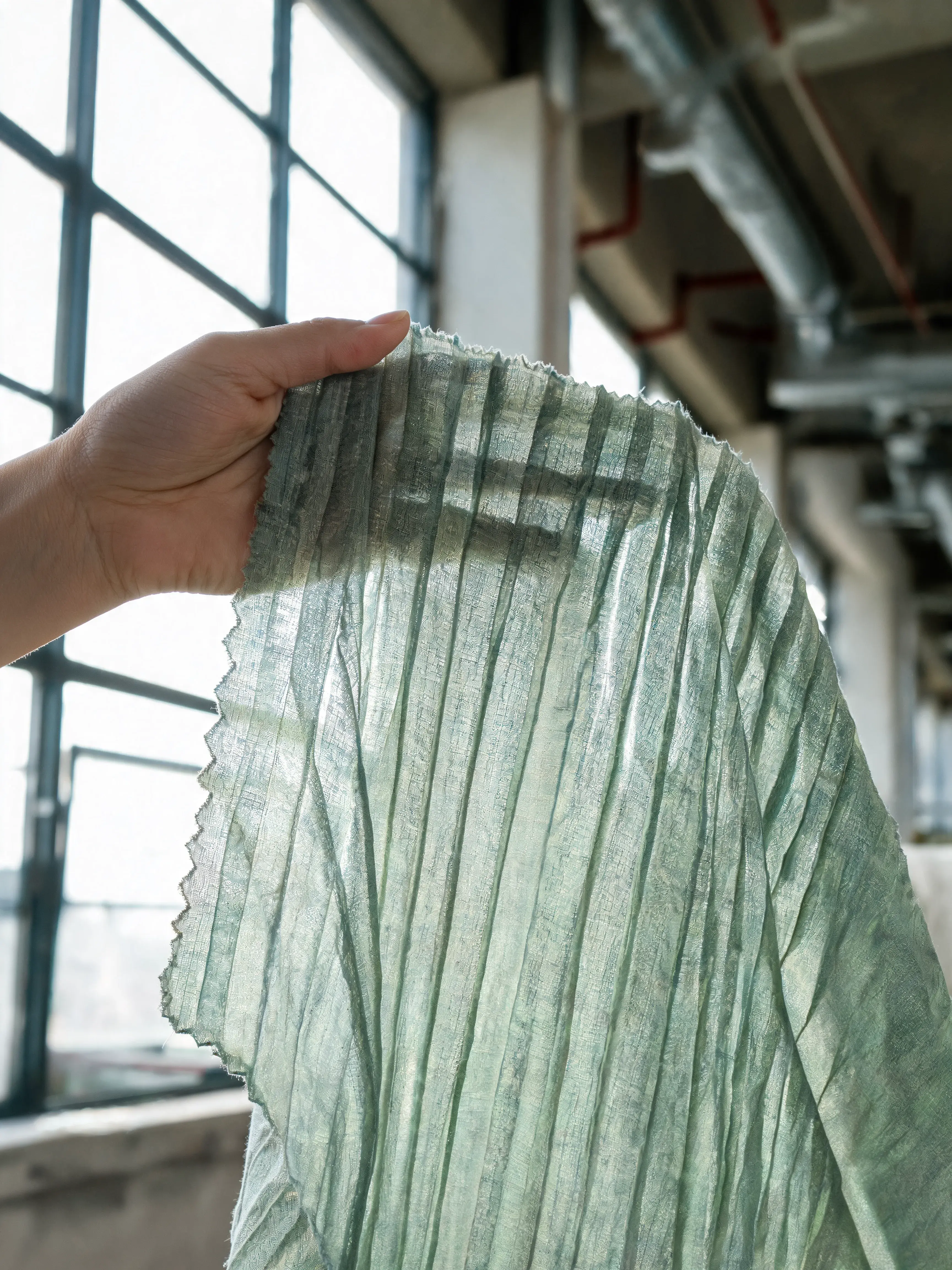 Main tenant tissu plissé vert sauge translucide – contre-jour, fenêtres industrielles, loft
