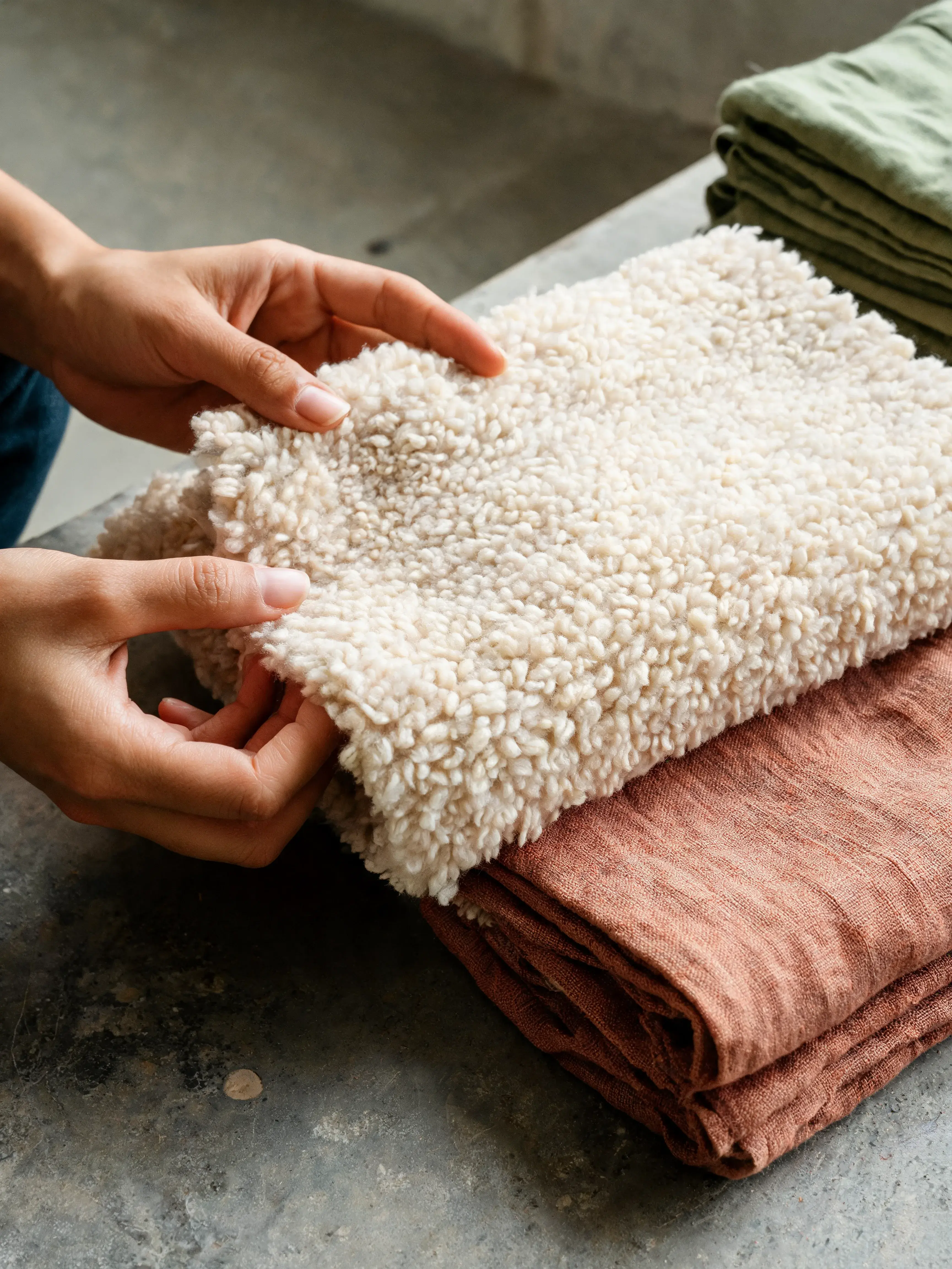 Mains évaluant texture pull bouclé crème – tissu lin terracotta dessous, sol béton, atelier mode