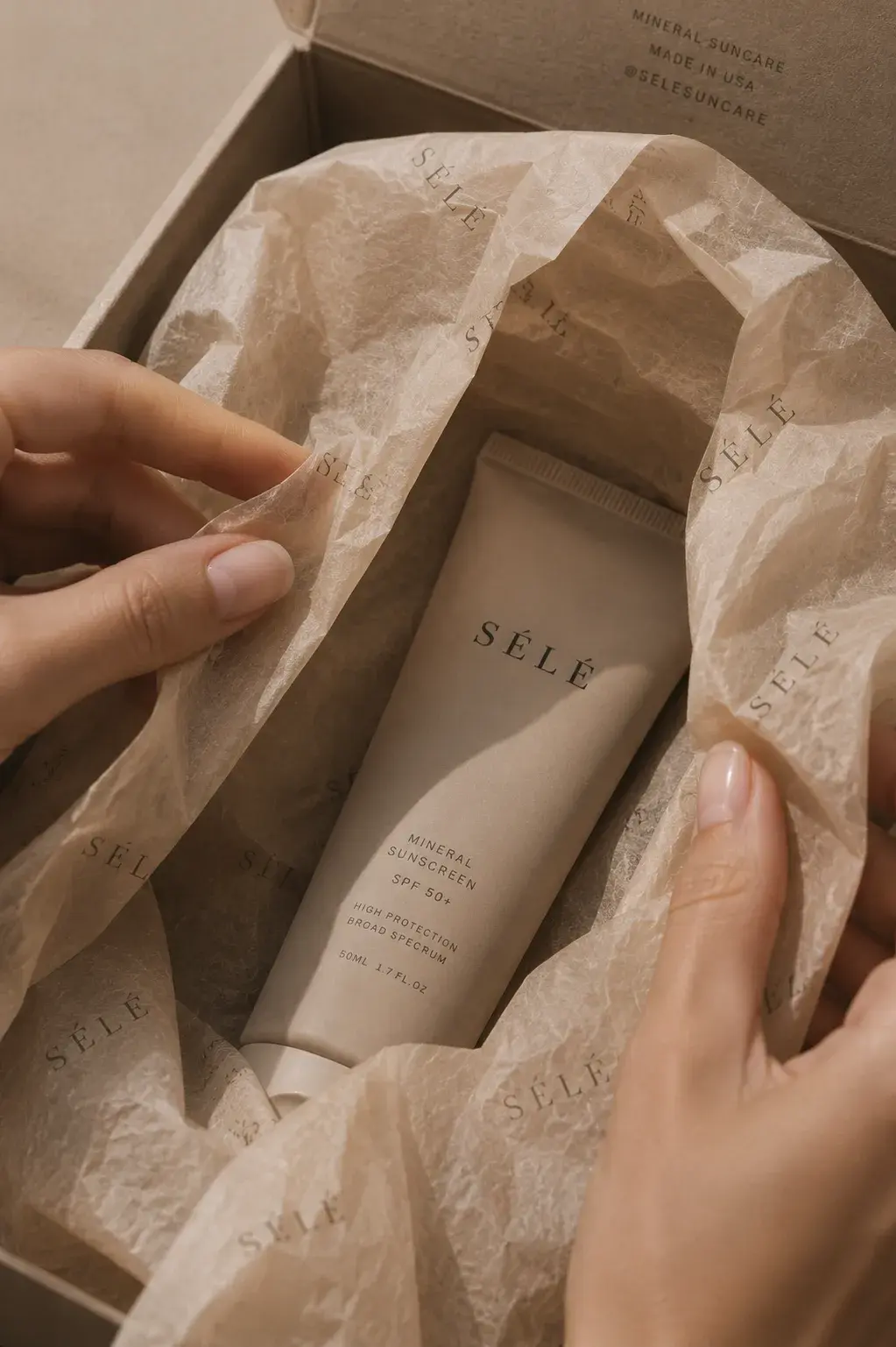 Mains féminines écartant papier de soie imprimé dans coffret – tube crème solaire, fond sombre