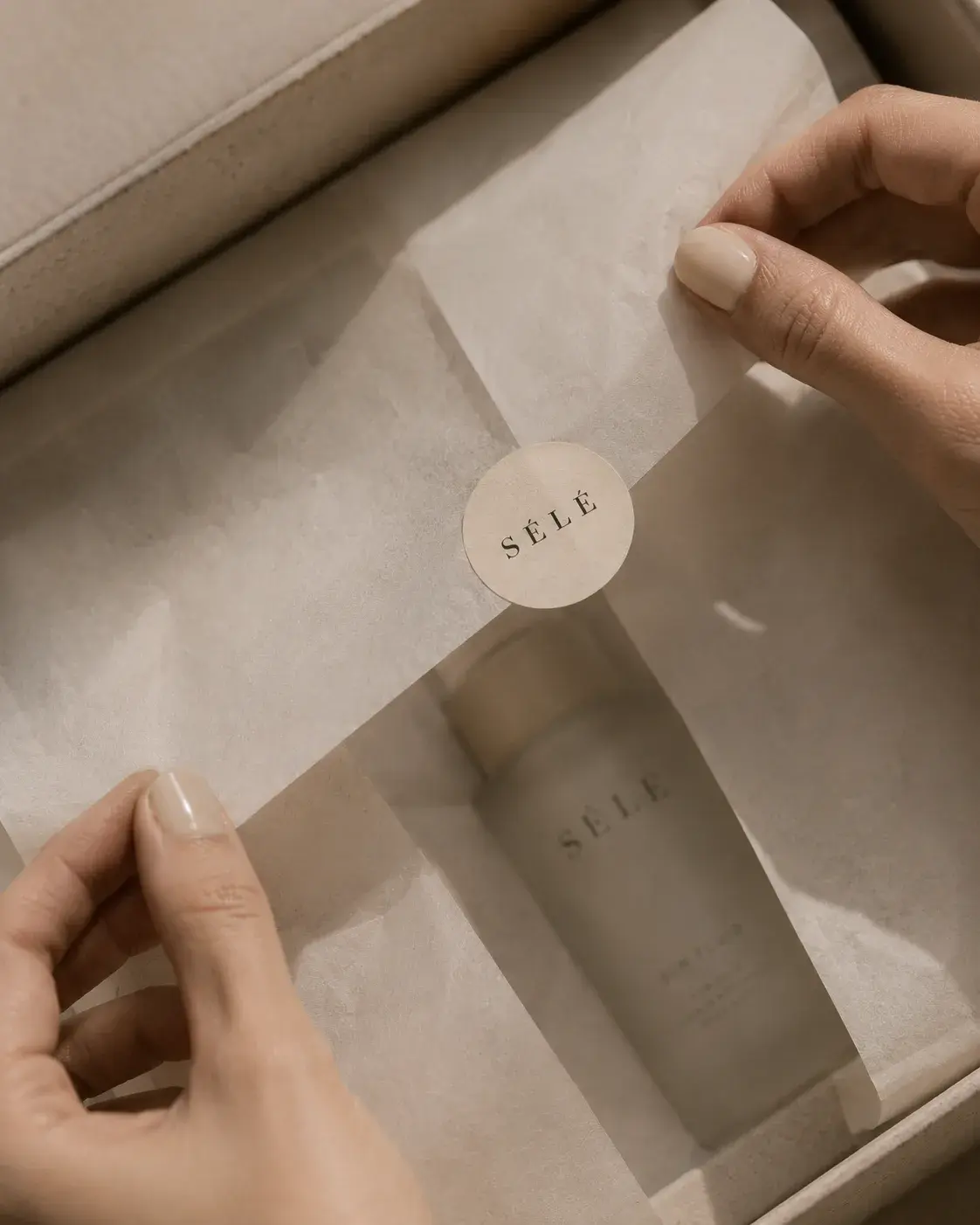 Mains féminines ouvrant un coffret cosmétique – sticker rond, tube soin, papier de soie, kraft