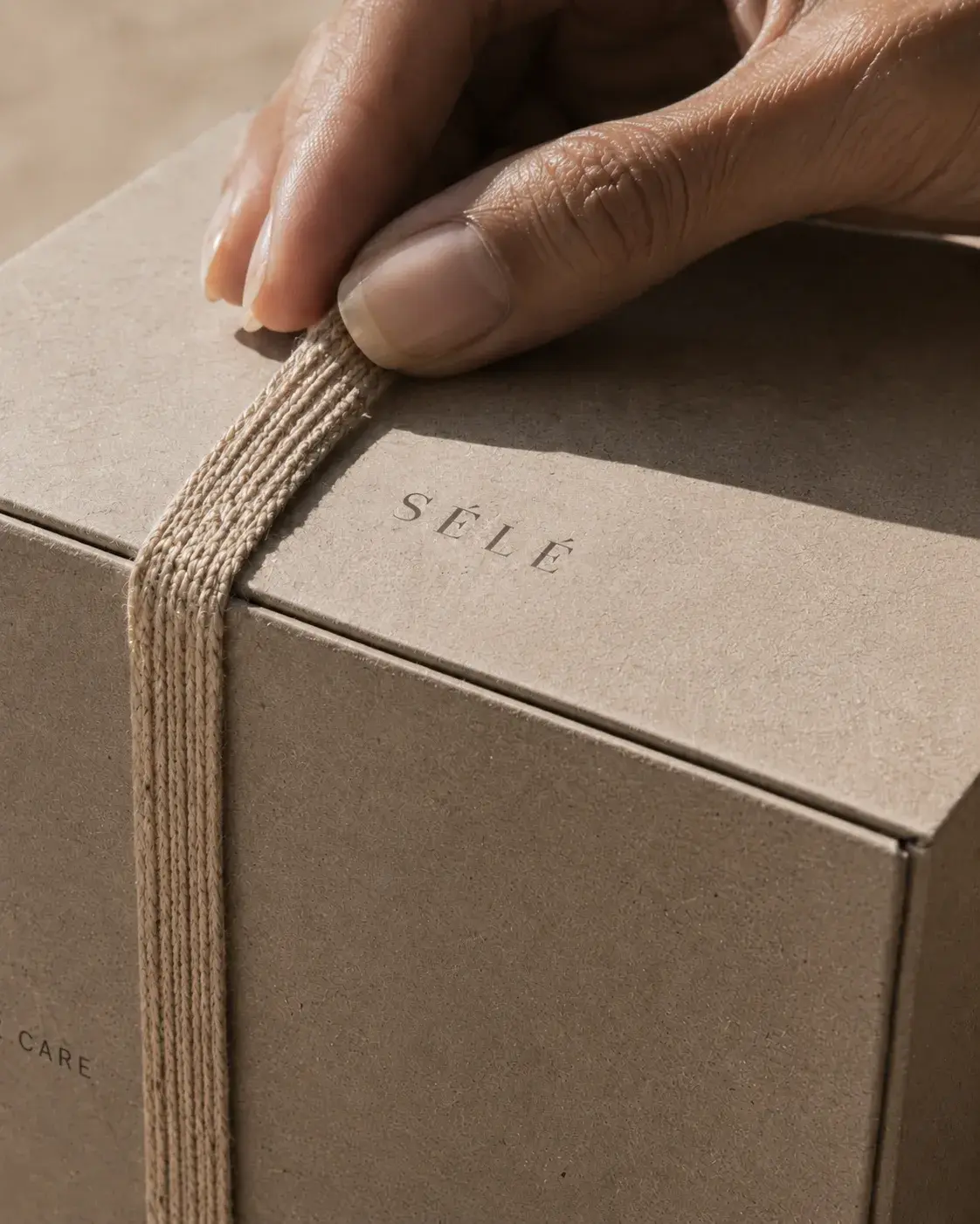 Mains peau noire posées sur coffret kraft noué avec ruban jute – sable, lumière rasante