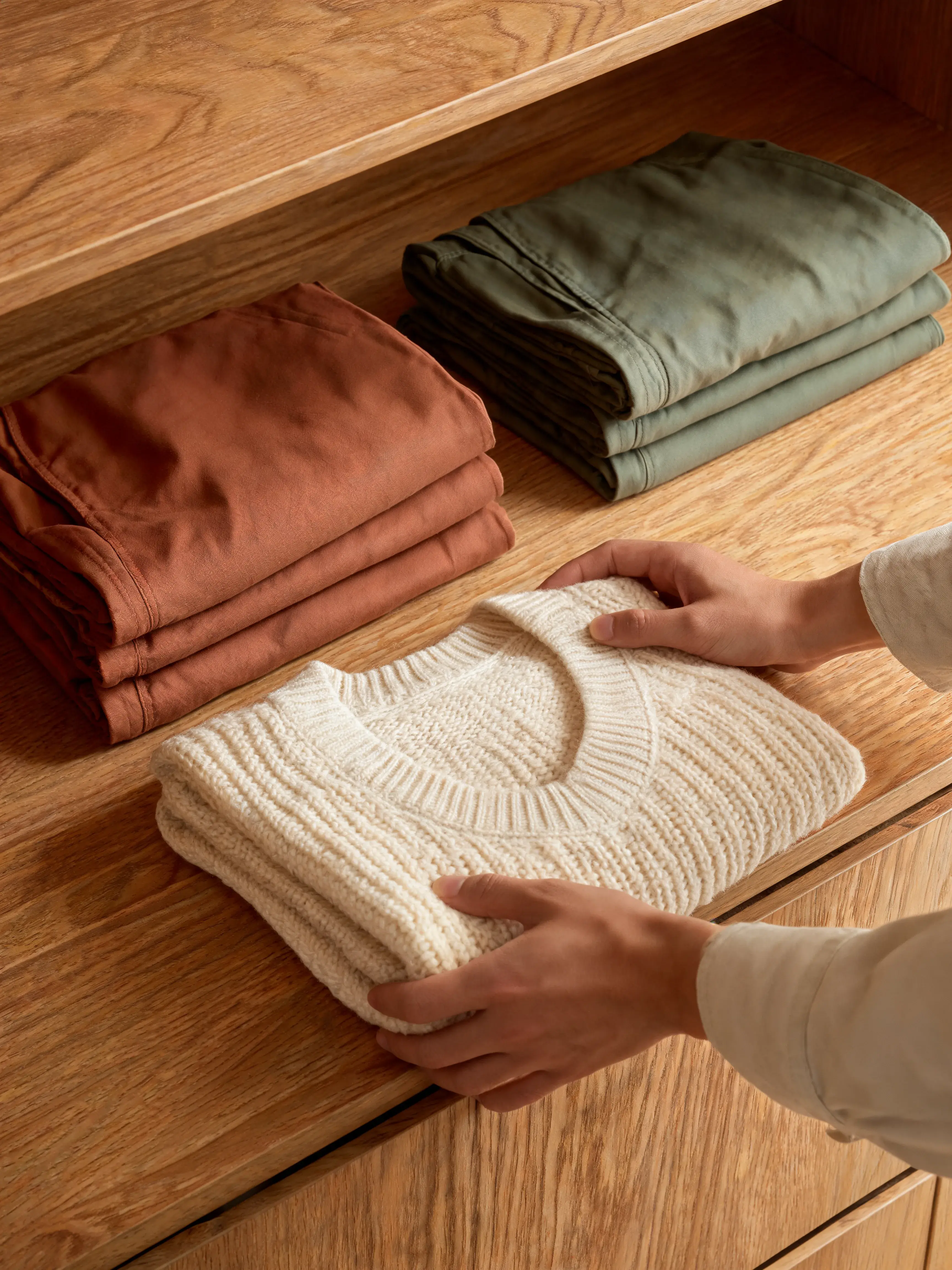 Mains rangeant pull tricoté crème – meuble bois tiroir, pantalons pliés terracotta et vert