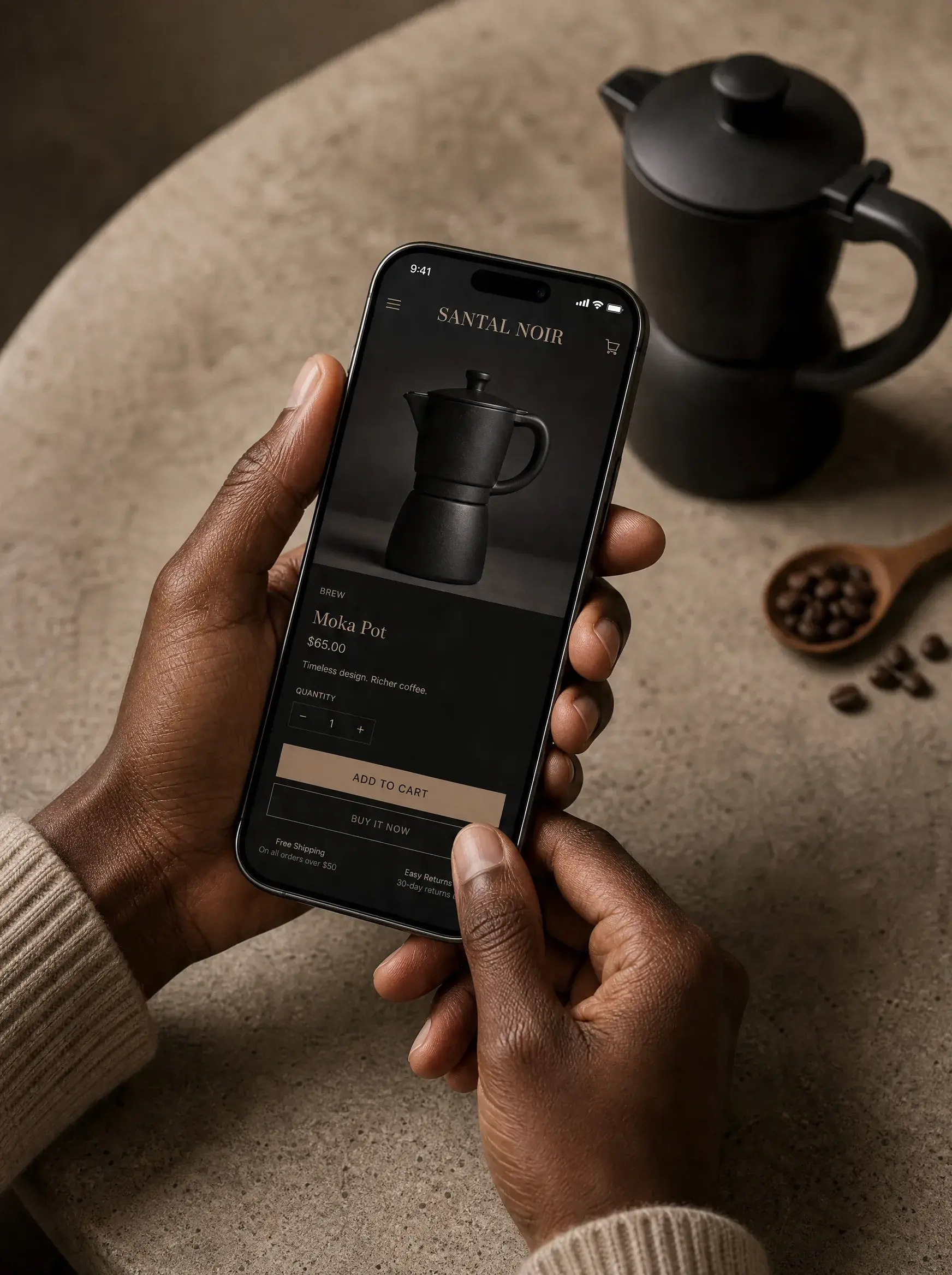 Mains tenant smartphone montrant page boutique cafetière moka – table béton, grains, cuillère bois