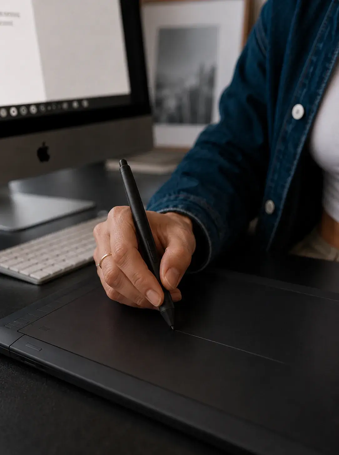 Mains tenant un stylet au-dessus d'une tablette graphique Wacom noire, iMac et vue urbaine en fond