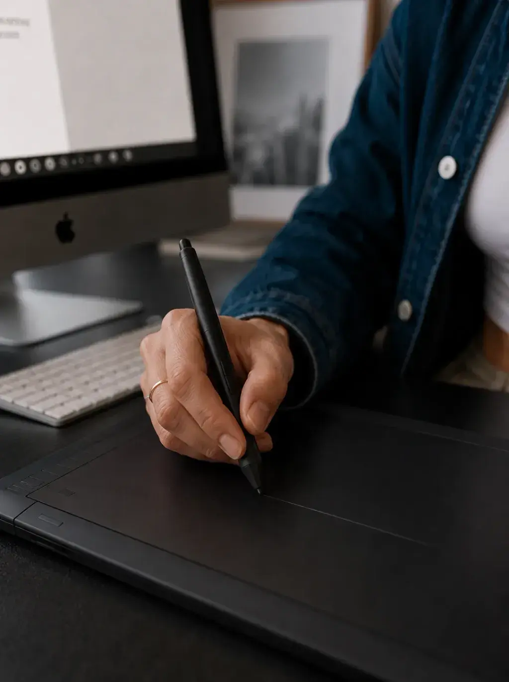 Mains tenant un stylet au-dessus d'une tablette graphique Wacom noire, iMac et vue urbaine en fond
