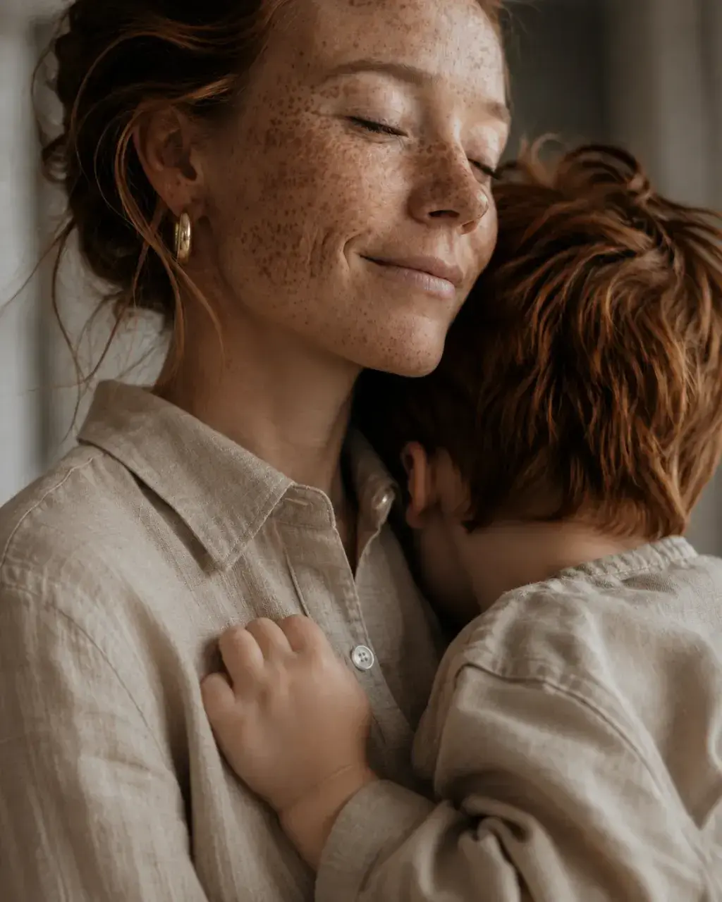 Mère aux taches de rousseur souriant les yeux fermés serrant son enfant roux – intérieur lin beige