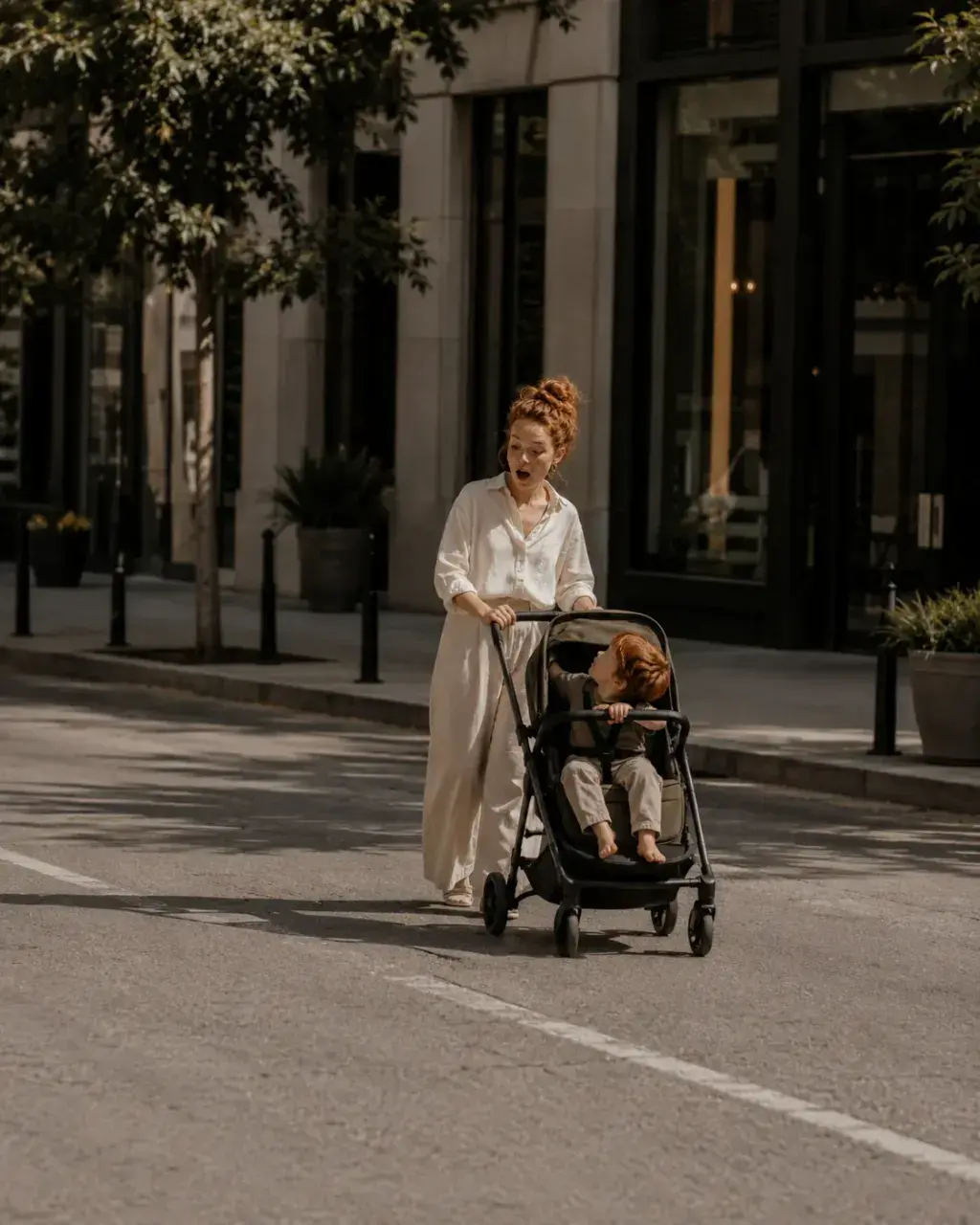 Mère en robe blanche poussant une poussette dans une avenue – enfant roux, lumière estivale
