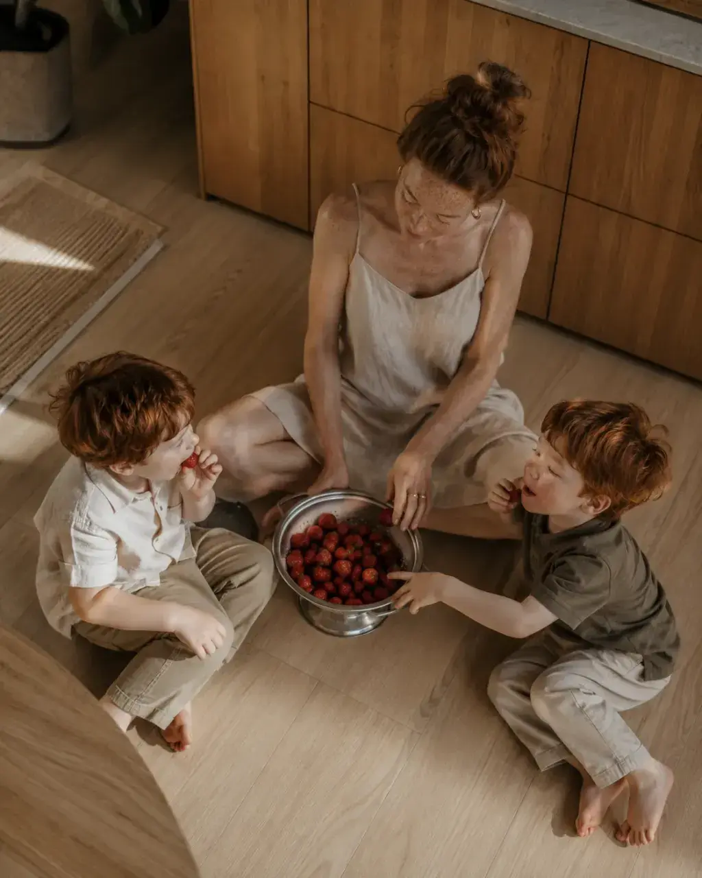 Mère et deux enfants roux mangeant des fraises par terre – cuisine en bois lumineuse