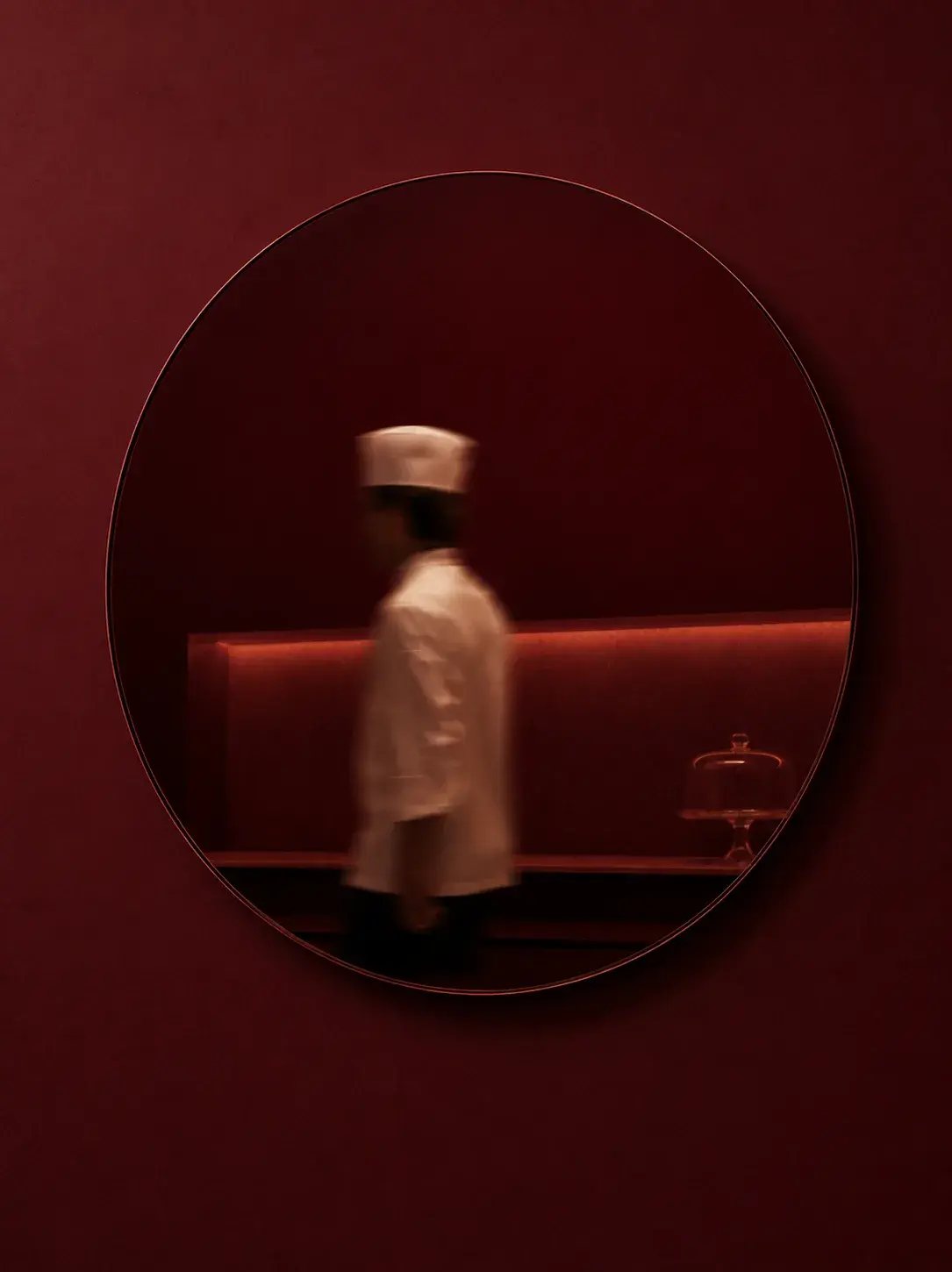 Miroir circulaire bordeaux sur mur bordeaux – reflet flou de cuisinier en toque passant en brasserie