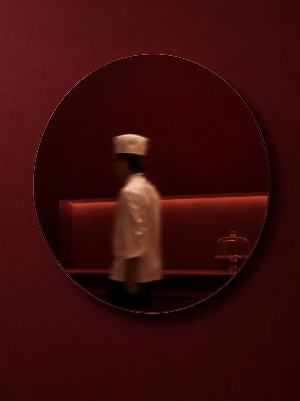 Miroir circulaire bordeaux sur mur bordeaux – reflet flou de cuisinier en toque passant en brasserie