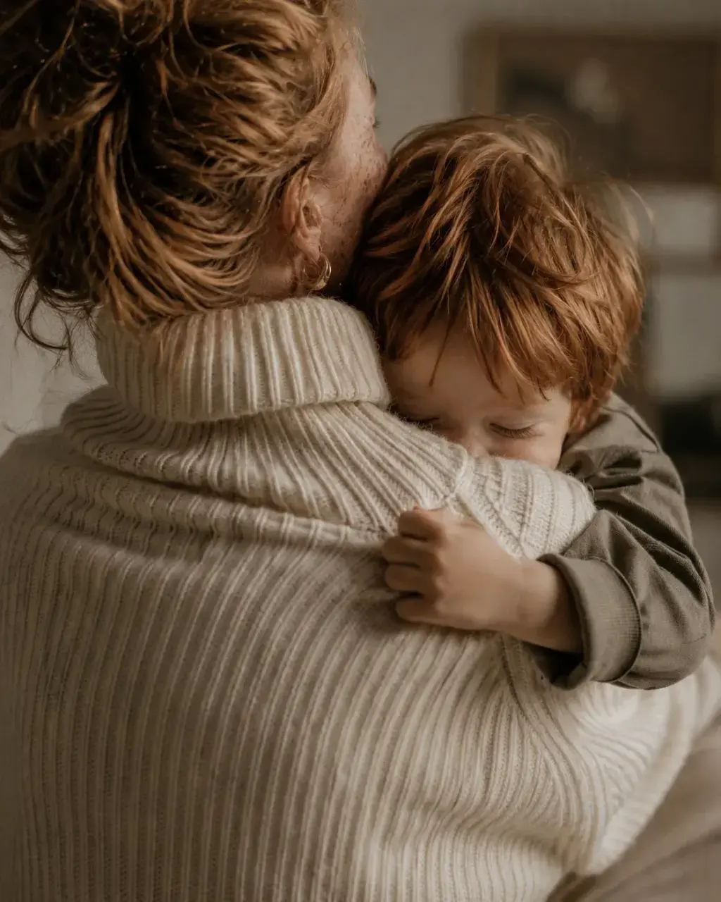 Parent en pull col roulé beige portant un enfant roux dans les bras – de dos, intérieur