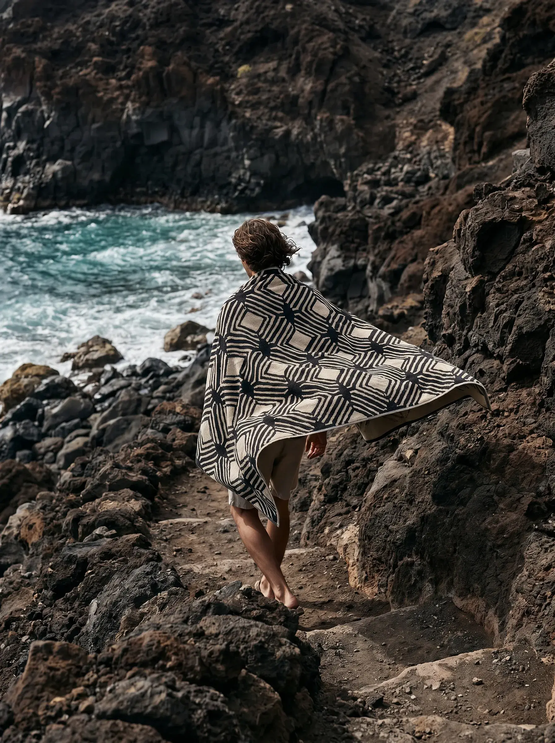 Personne enveloppée d'un poncho à carreaux géométriques sur falaises rocheuses face à l'océan