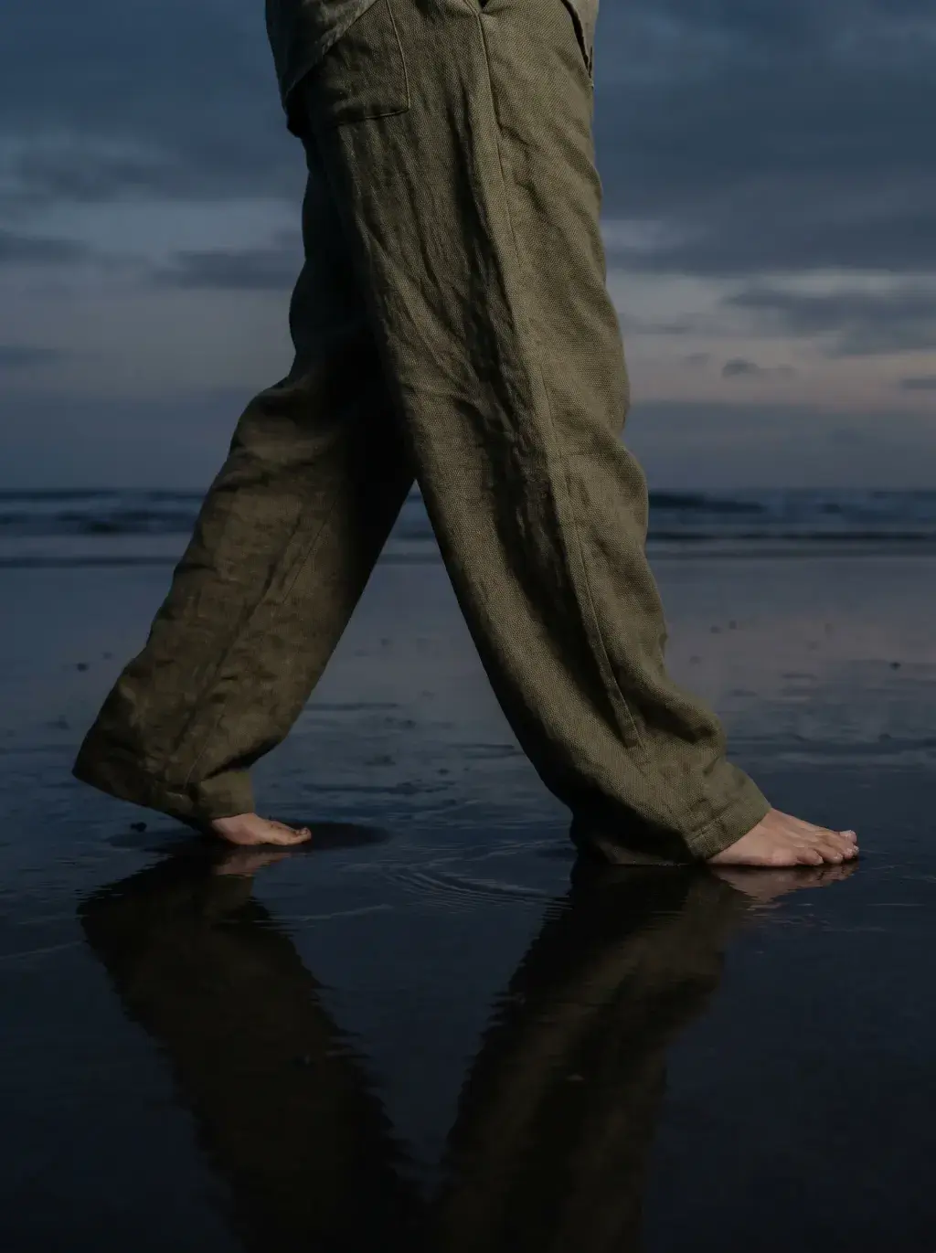 Pieds nus et bas de pantalon kaki en lin sur sable volcanique noir mouillé